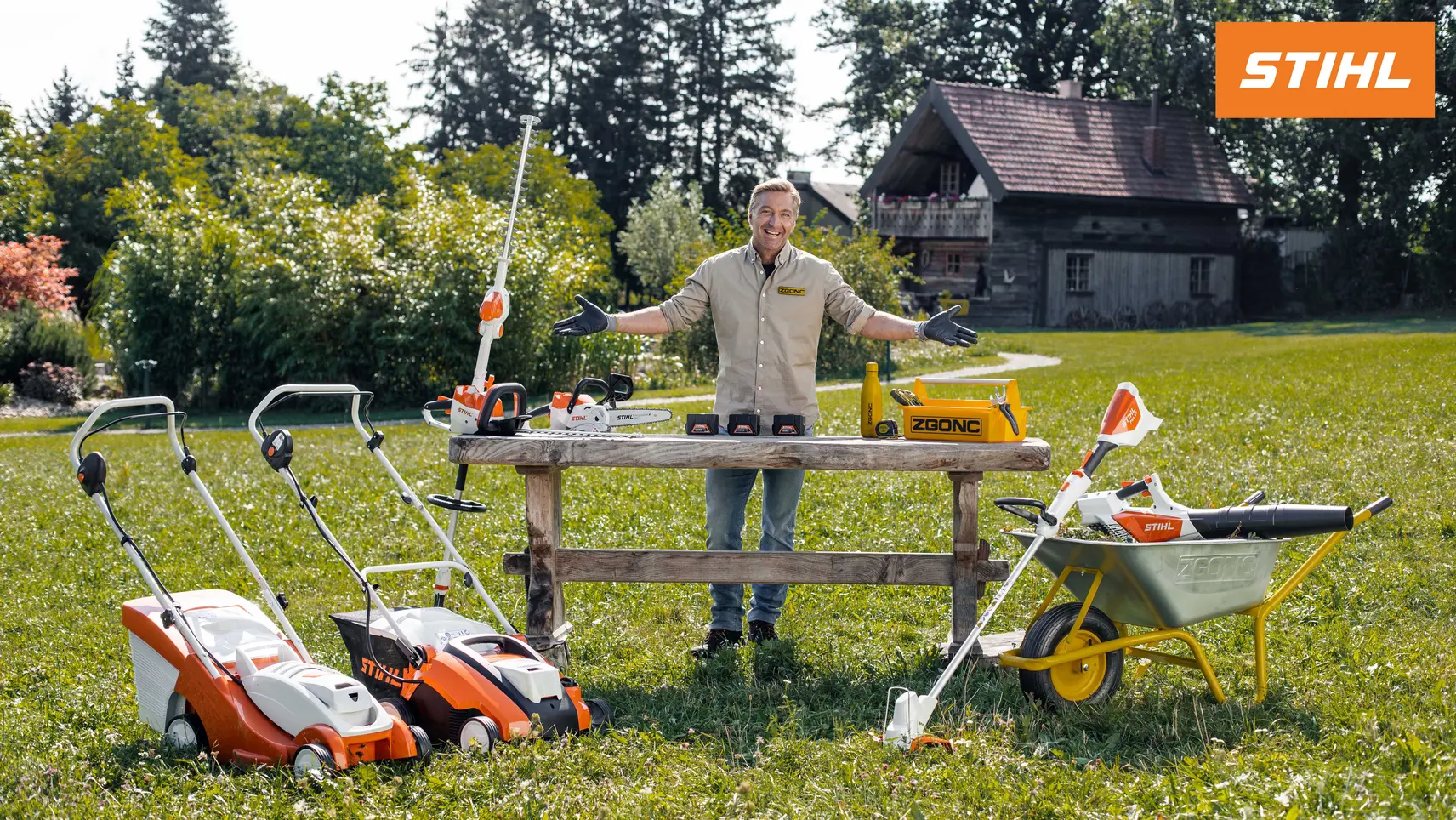 Mann präsentiert STIHL Gartengeräte im Freien: Rasenmäher, Kettensägen, Heckenschere und Schubkarre vor einem Landhaus.
