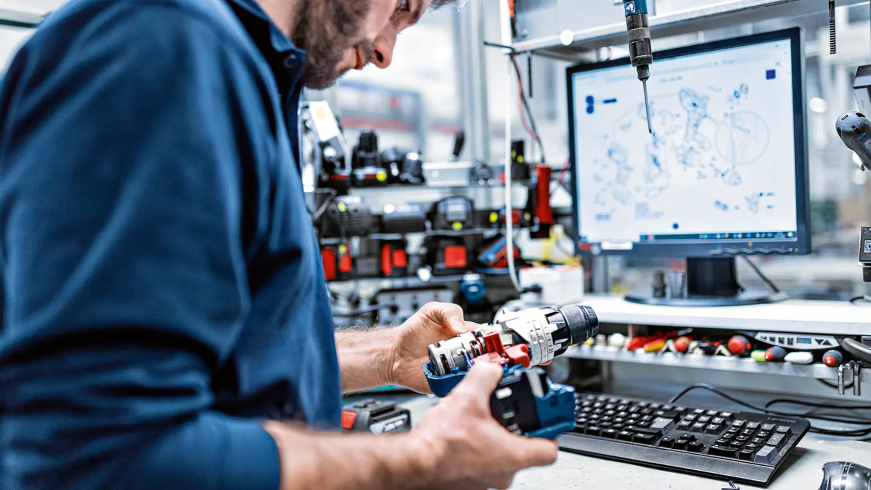 Techniker untersucht einen Akkubohrschrauber in einer Werkstatt, im Hintergrund Monitor mit technischer Zeichnung und Elektrowerkzeuge.