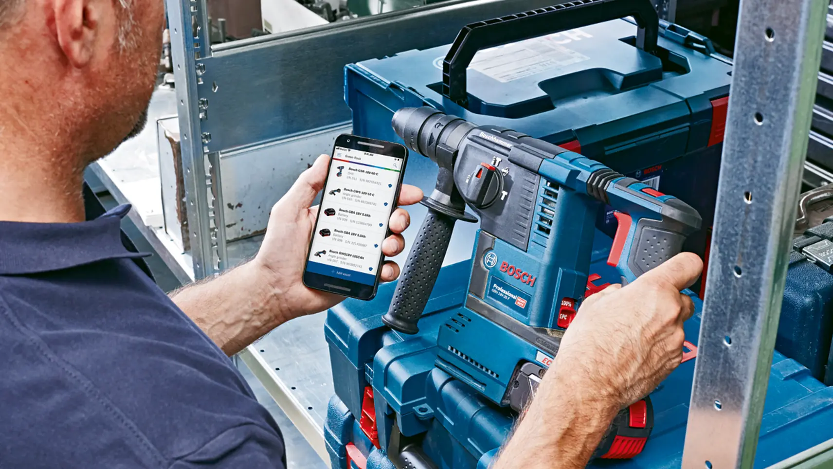 Mann hält Smartphone mit Bosch-App neben einem Bosch Professional Bohrhammer in blauem Werkzeugkoffer im Regal.
