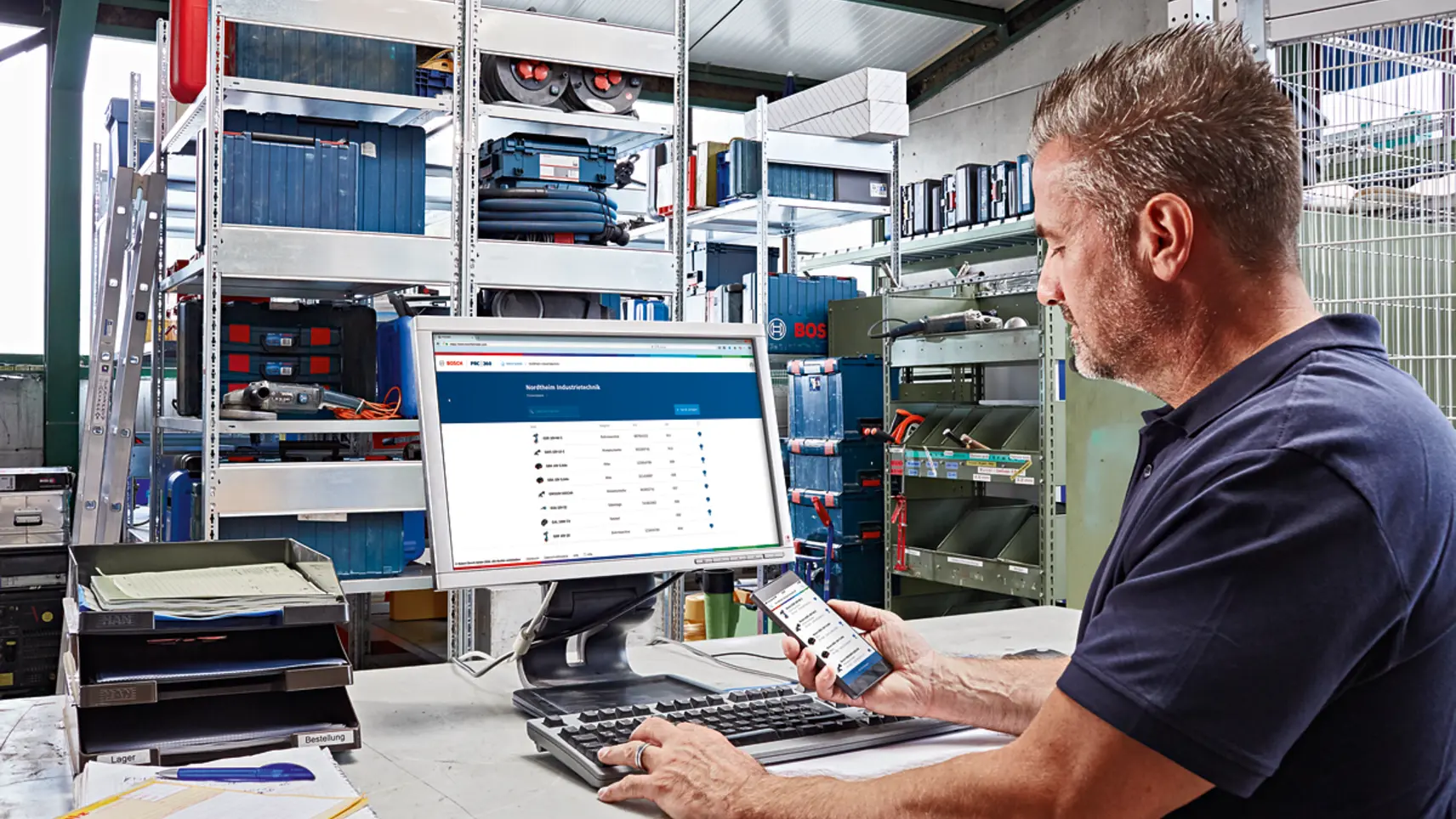 Mann in Werkstatt arbeitet am PC mit Bosch-Software, hält Smartphone in der Hand, Werkzeugregale im Hintergrund.