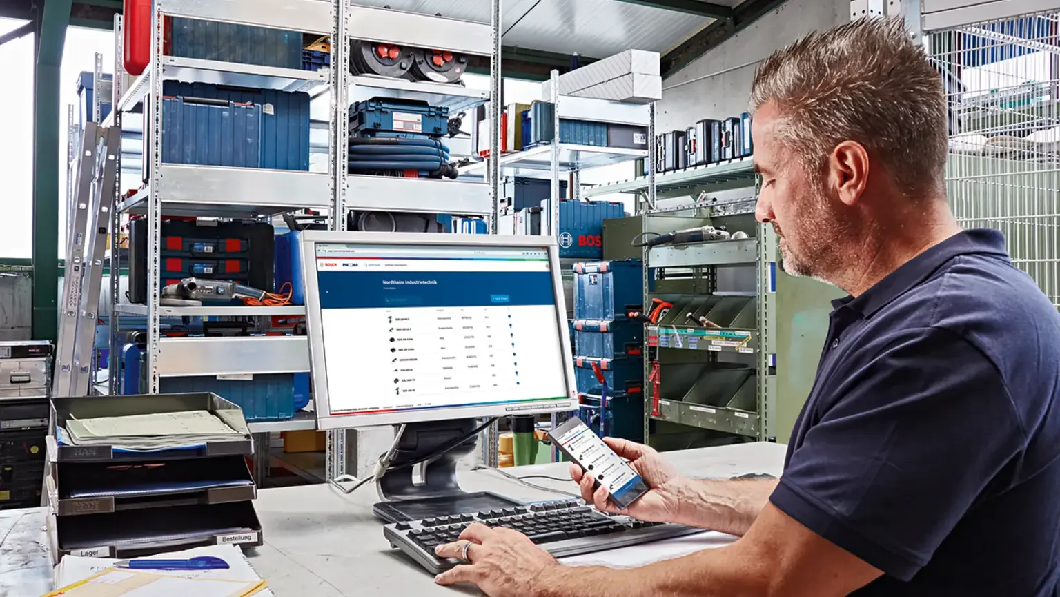 Mann in Werkstatt arbeitet am PC mit Bosch-Software, hält Smartphone in der Hand, Werkzeugregale im Hintergrund.
