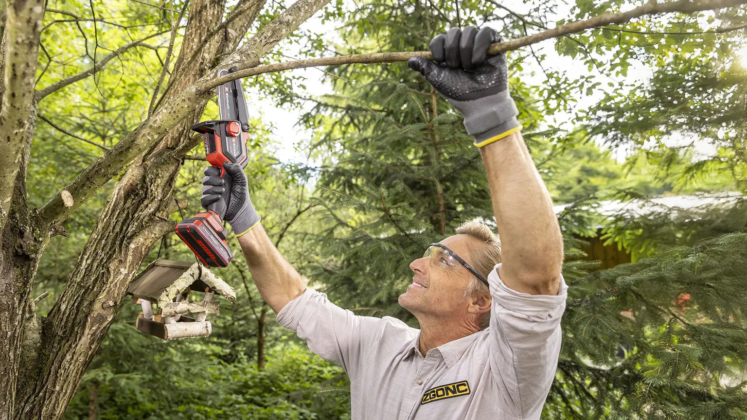 Mann mit Schutzbrille und Handschuhen sägt mit einer Einhell Akku-Säge einen Ast im Garten ab.