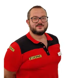 Mann mit Brille und Bart trägt ein rot-schwarzes ZGONC-Poloshirt mit Namensschild, Porträtfoto vor weißem Hintergrund.