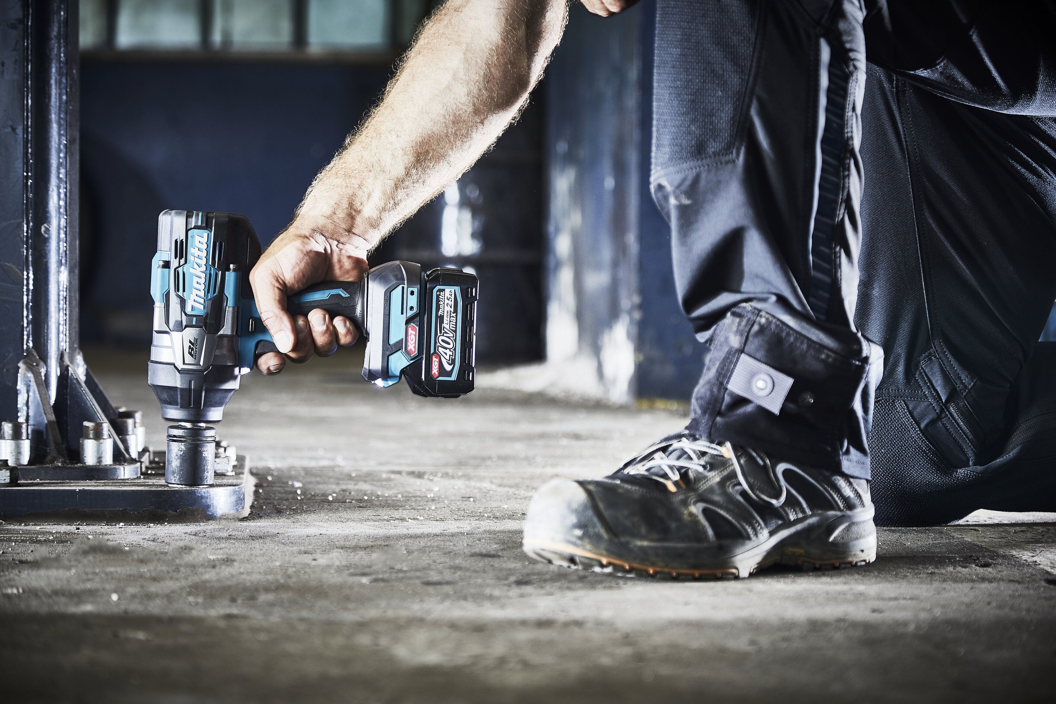 Handwerker kniet auf Betonboden und verwendet einen blauen Makita 40V XGT Akku-Schlagschrauber auf Metallbefestigung.