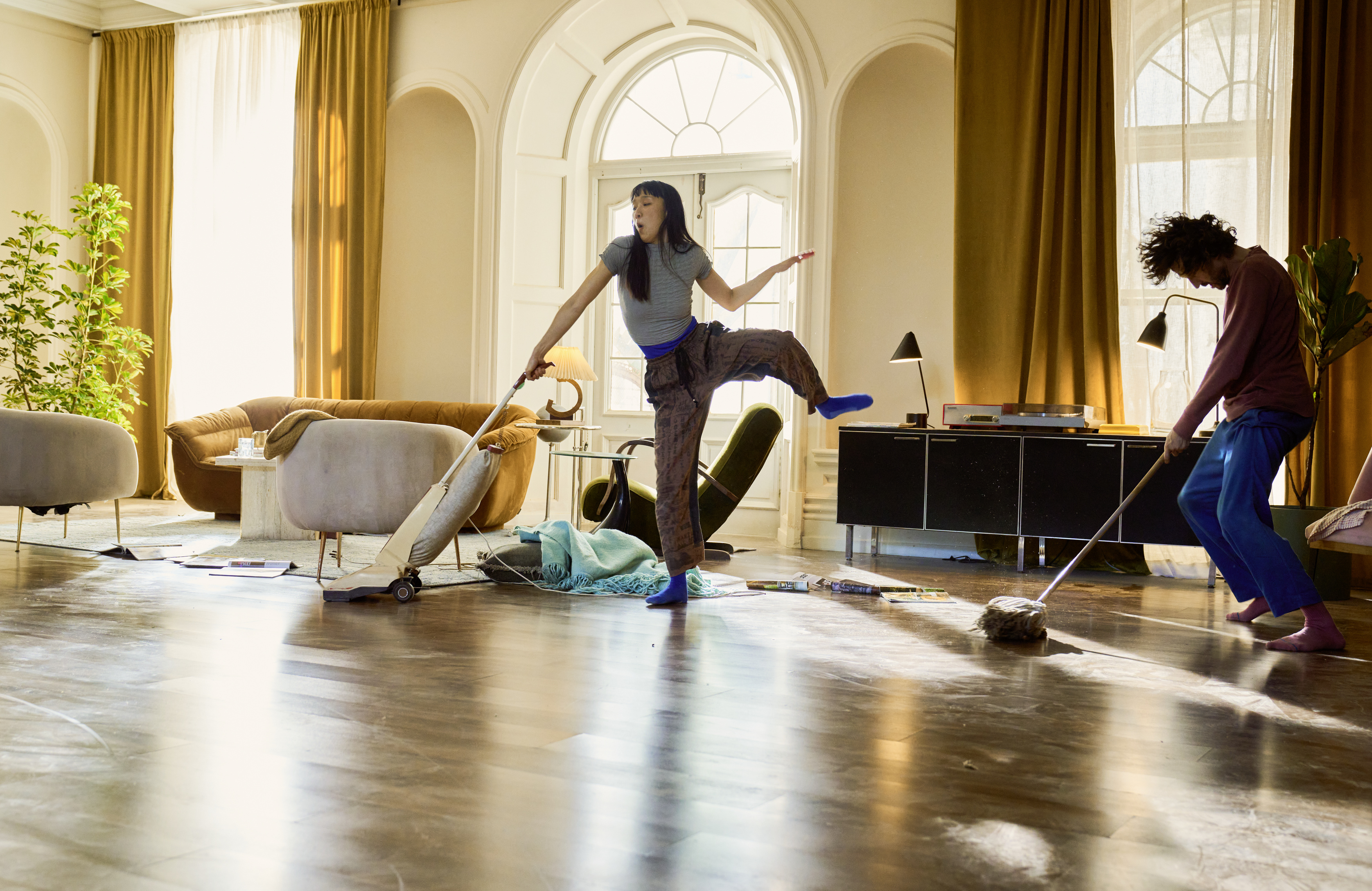 Zwei Personen putzen ein sonnendurchflutetes Wohnzimmer; die eine staubsaugt in einer verspielten Pose, die andere wischt den Boden. Im Hintergrund stehen gemütliche Möbel und Pflanzen.
