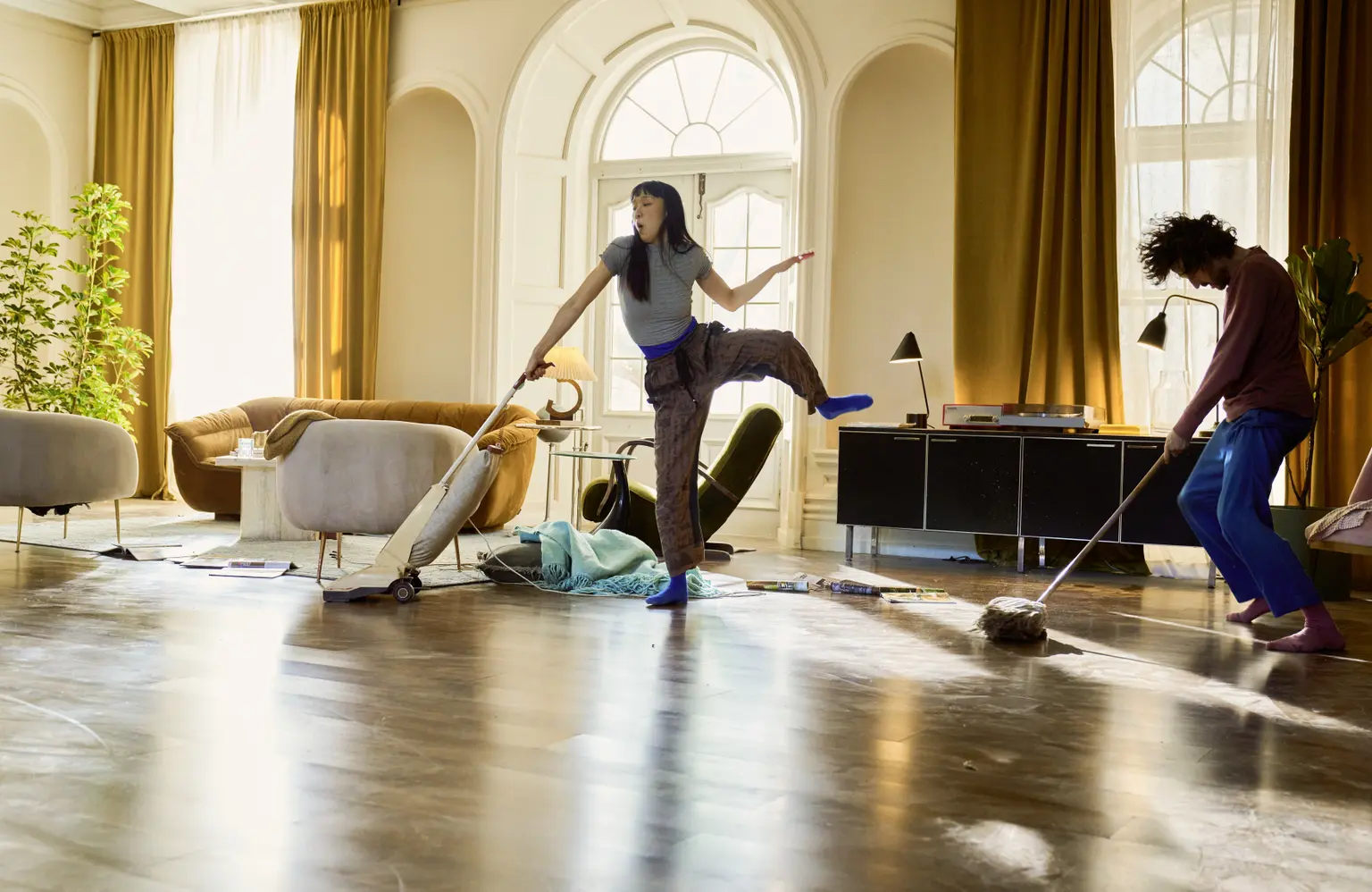 Zwei Personen putzen ein sonnendurchflutetes Wohnzimmer; die eine staubsaugt in einer verspielten Pose, die andere wischt den Boden. Im Hintergrund stehen gemütliche Möbel und Pflanzen.
