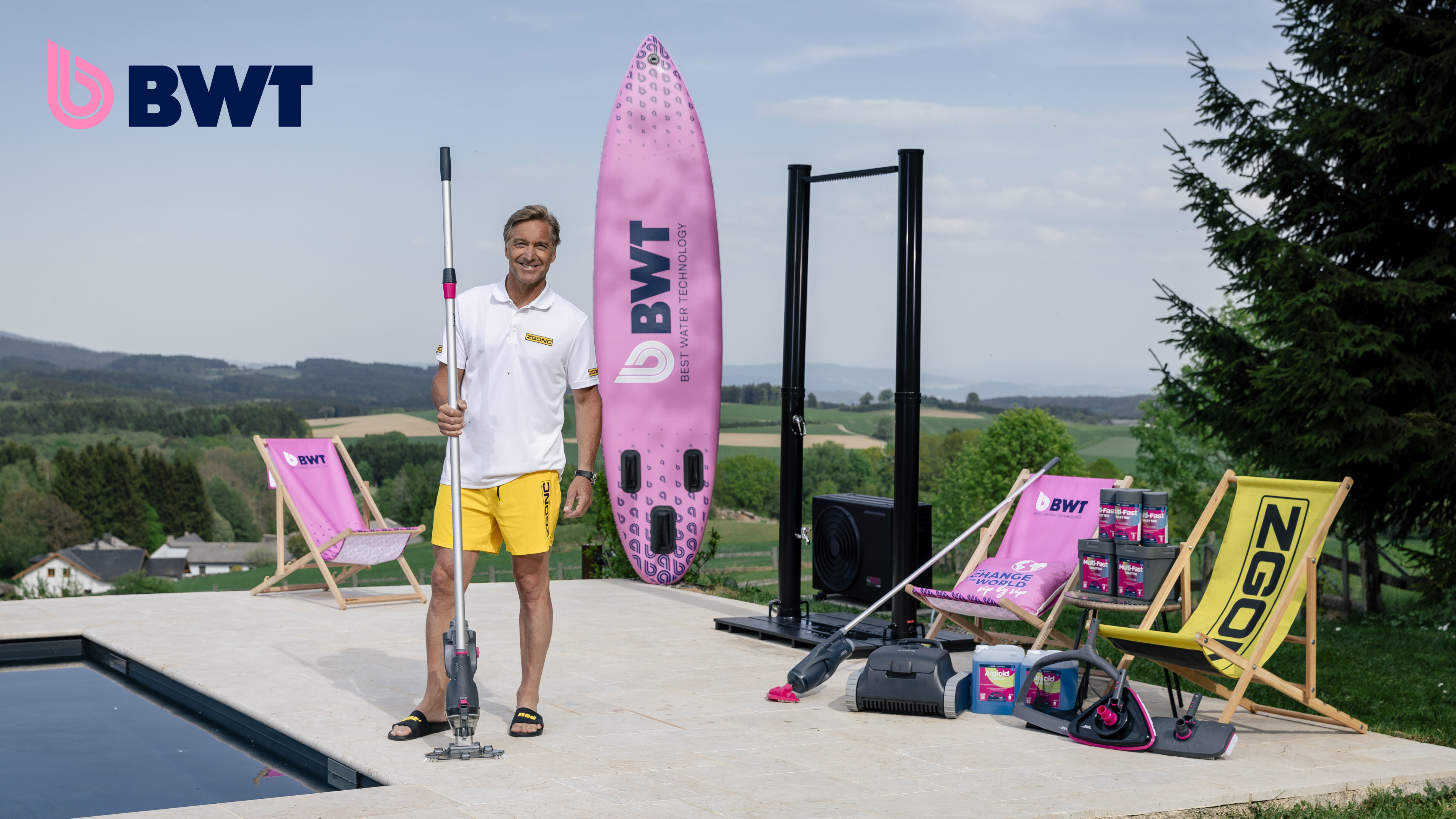 Mann mit BWT-Poolreiniger neben pinkem BWT-SUP-Board, Liegestühlen und Poolzubehör auf einer Terrasse mit Panoramablick.