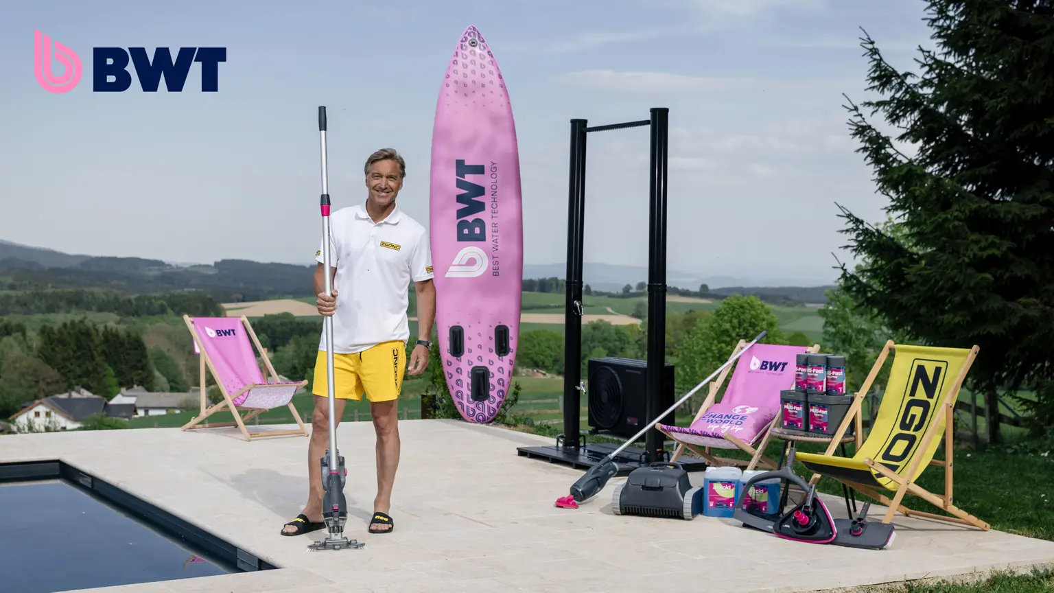 Mann mit BWT-Poolreiniger neben pinkem BWT-SUP-Board, Liegestühlen und Poolzubehör auf einer Terrasse mit Panoramablick.