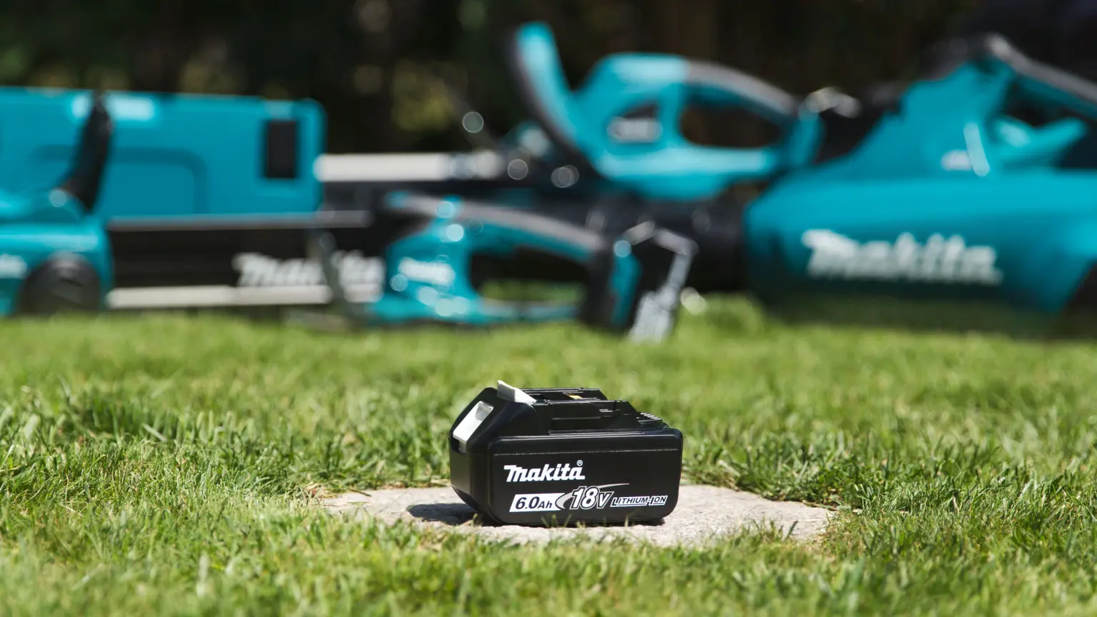 Makita 18V 6Ah Lithium-Ionen-Akku auf Rasen, im Hintergrund türkisfarbene Makita-Gartengeräte unscharf.