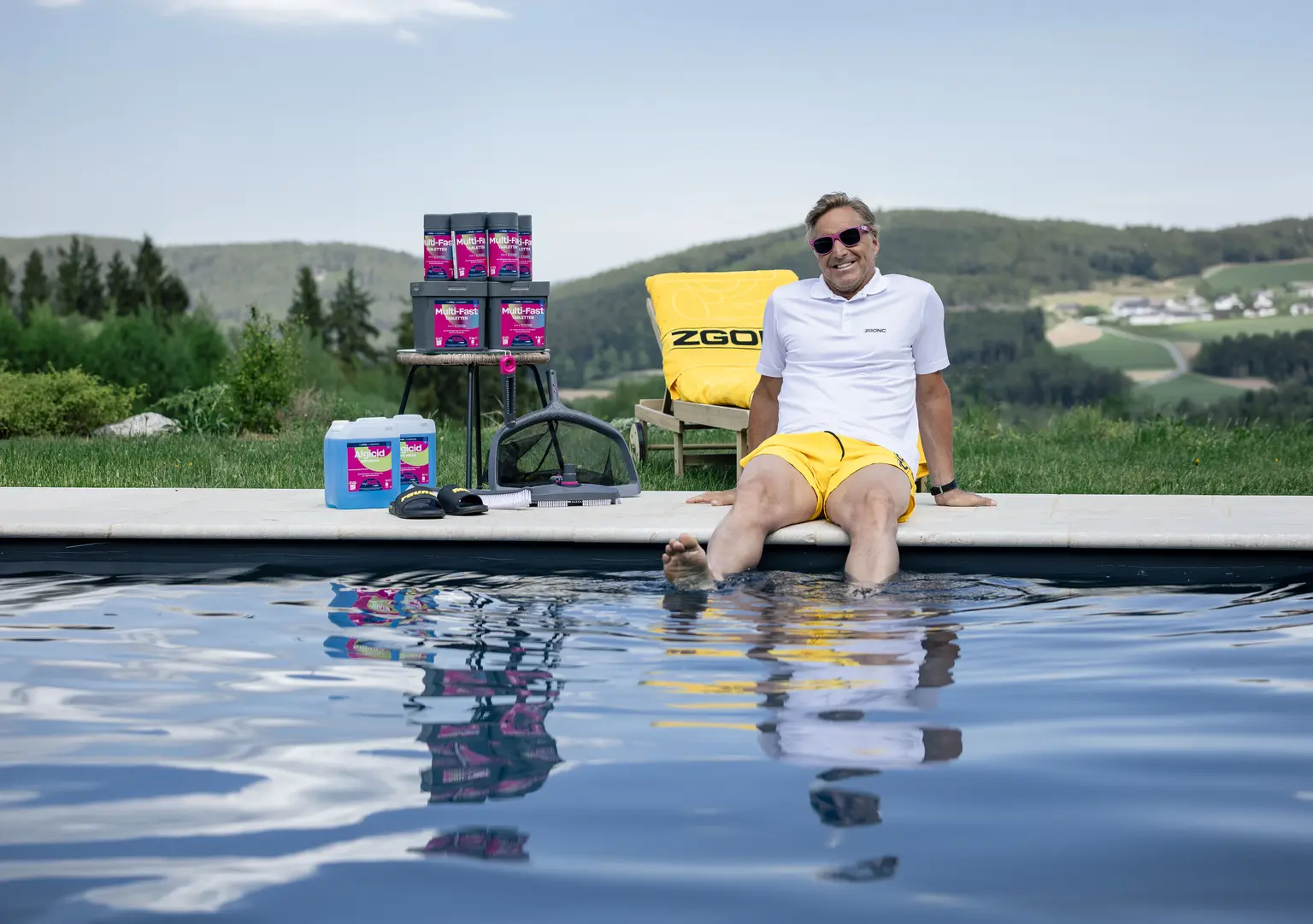 Mann mit Sonnenbrille sitzt am Poolrand mit Füßen im Wasser, neben Poolpflegemitteln der Marke Multi-Fast, grüne Berglandschaft im Hintergrund.
