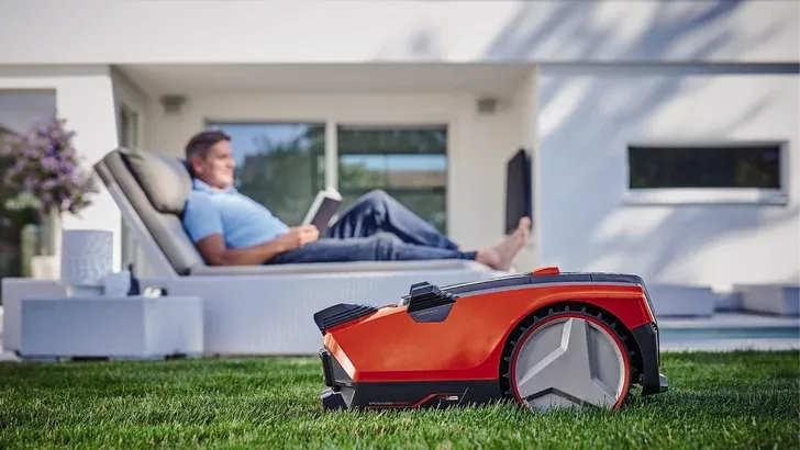 Ein Rasenmähroboter mäht den Rasen, während ein Mann auf einem Terrassenstuhl vor einem modernen weißen Haus sitzt und ein Buch liest.