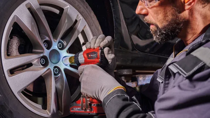Ein Mechaniker, der mit einem Elektrowerkzeug eine Radmutter an einem Auto festzieht und dabei Handschuhe und eine Schutzbrille trägt.
