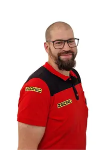 Mann mit Bart und Brille lächelt, trägt rot-schwarzes ZGONC Poloshirt, Porträtfoto vor weißem Hintergrund