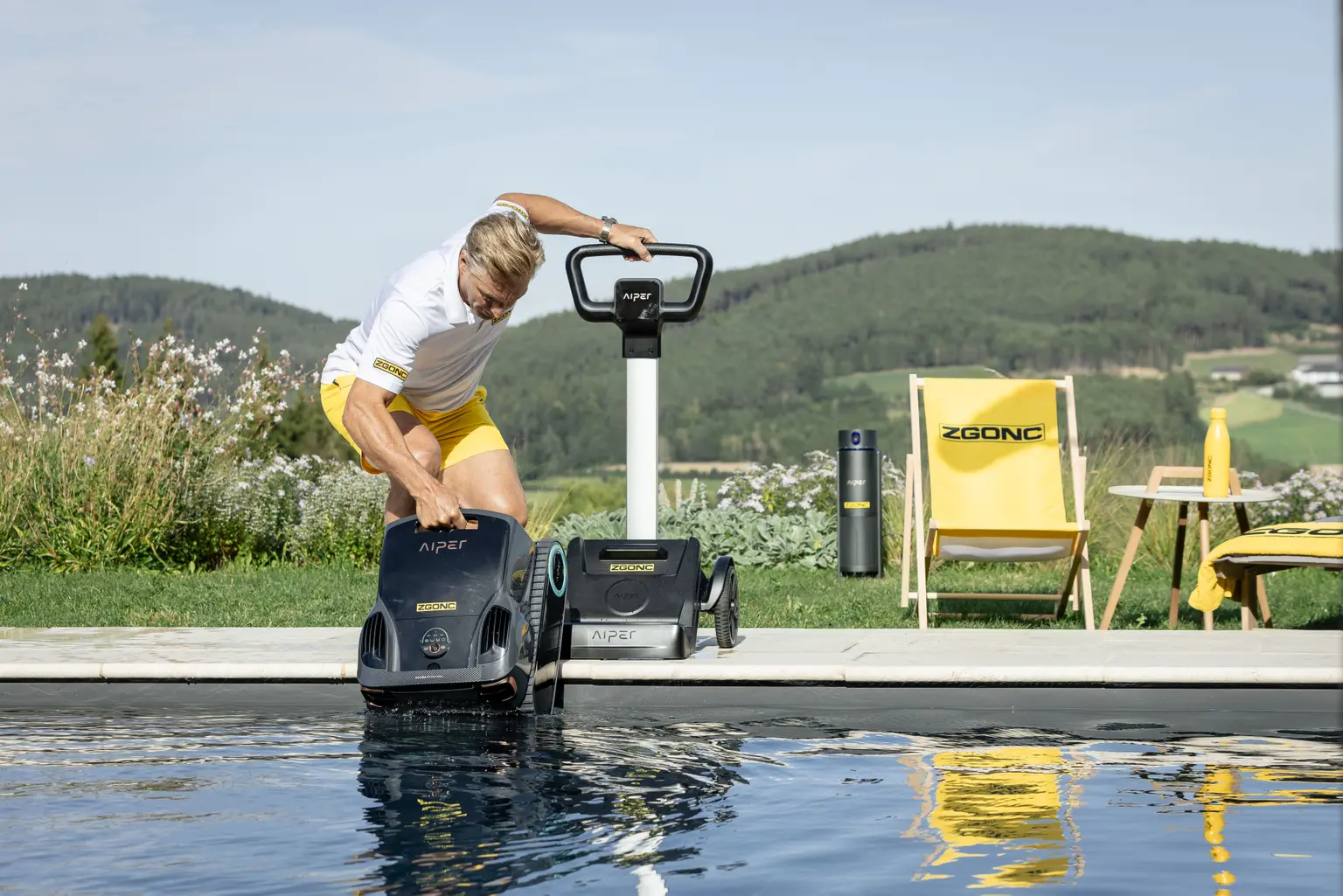 Mann setzt Aiper Poolroboter mit ZGONC-Branding in einen Außenpool, im Hintergrund Liegestuhl und grüne Hügel.