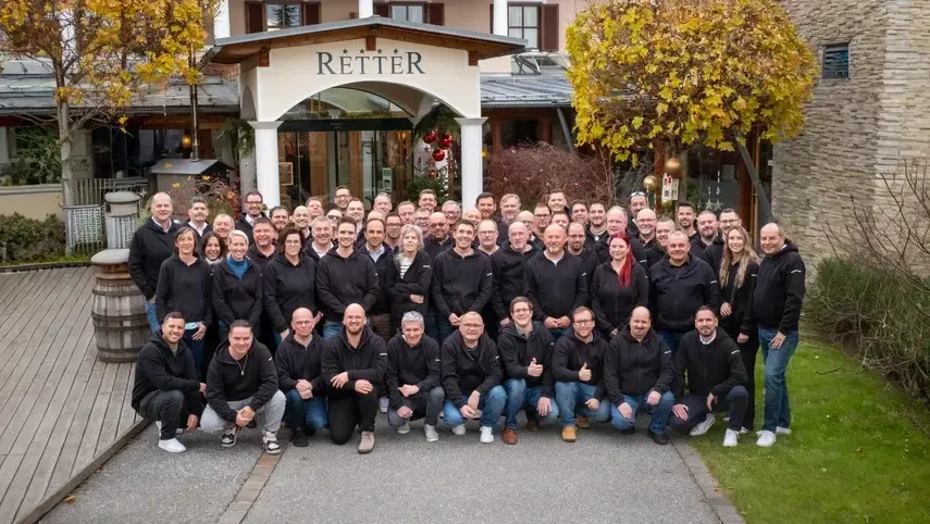 A large group of people in black jackets smiling and posing in front of a building with "RETTER" sign, surrounded by autumn trees.