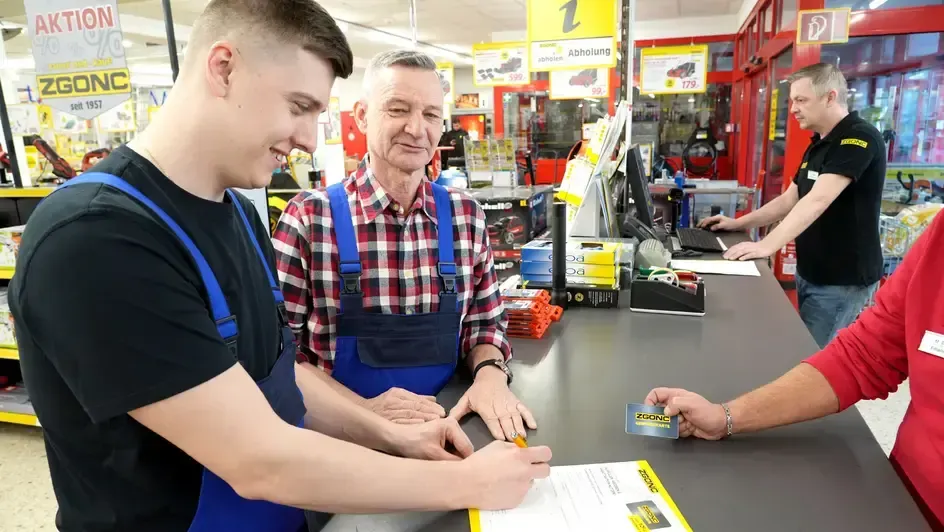 Zwei Mitarbeiter im ZGONC-Baumarkt an der Kasse, einer unterschreibt ein Dokument, Kassierer reicht eine Kundenkarte.