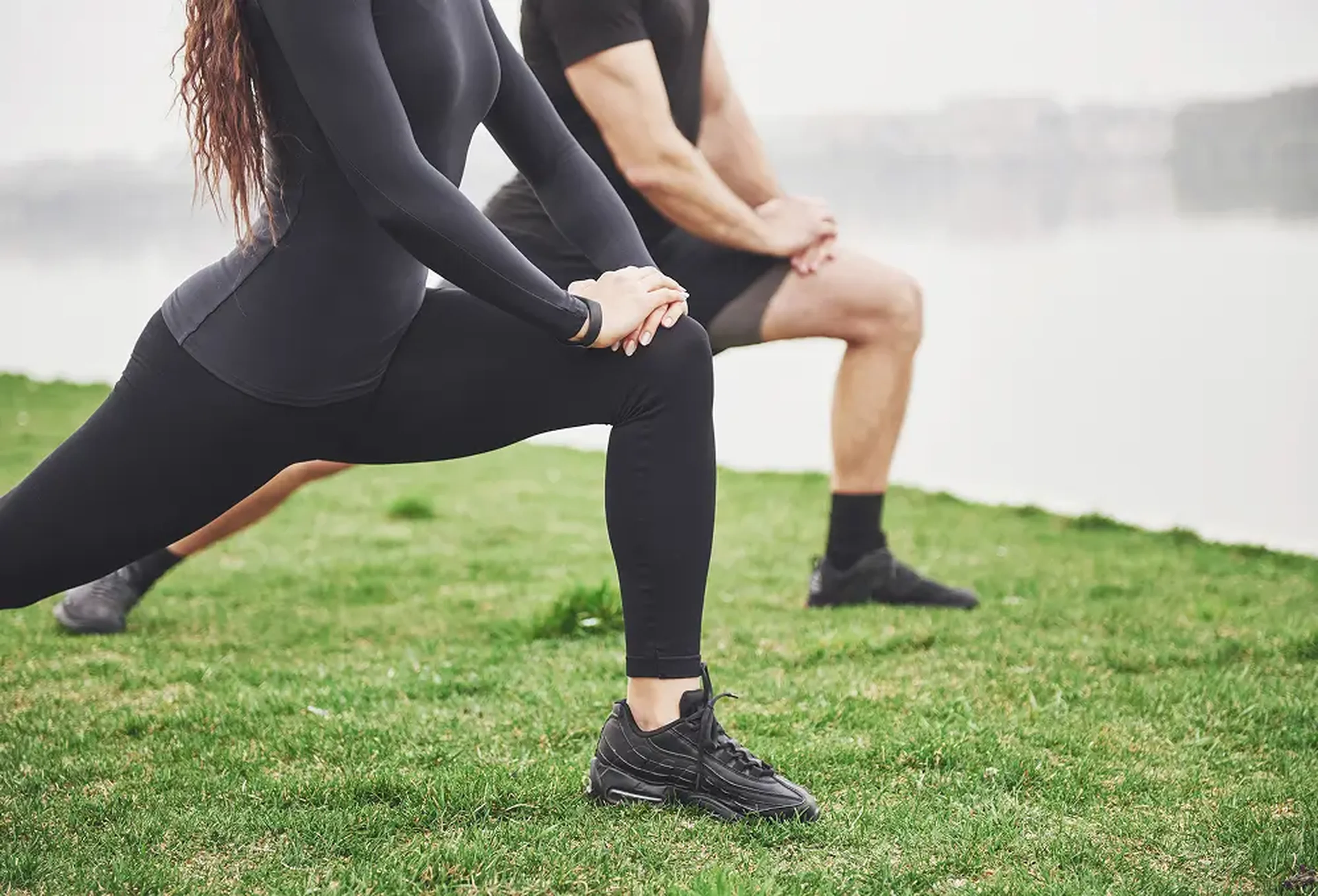 Two people in black athletic wear doing lunges together on grass near waterfront, demonstrating partner workout routine