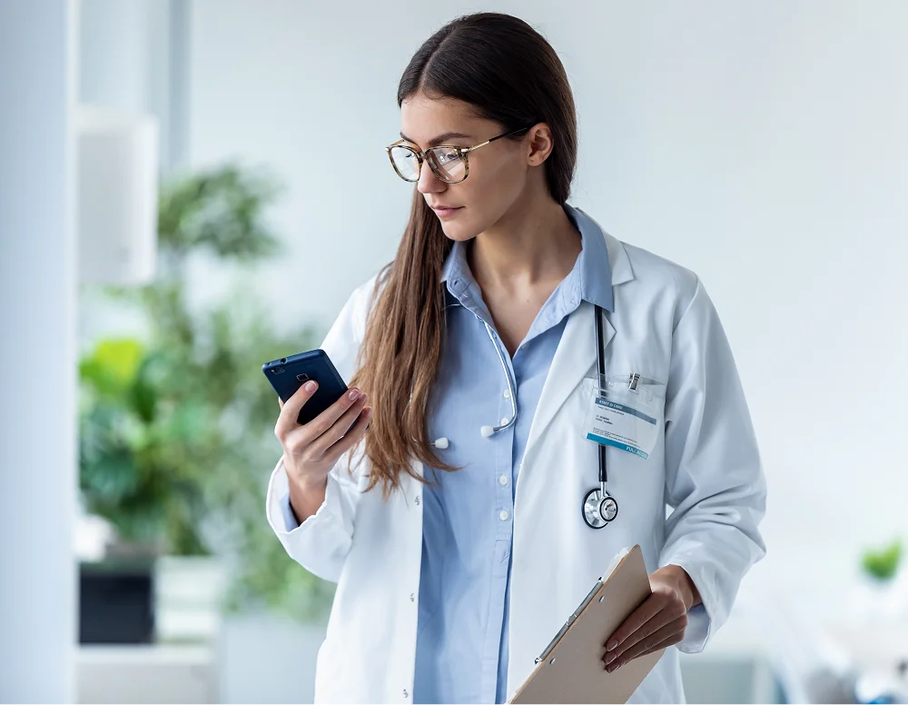 A young female doctor wearing glasses, white coat, and stethoscope looks at her smartphone while holding a clipboard in a bright medical office with plants in the background