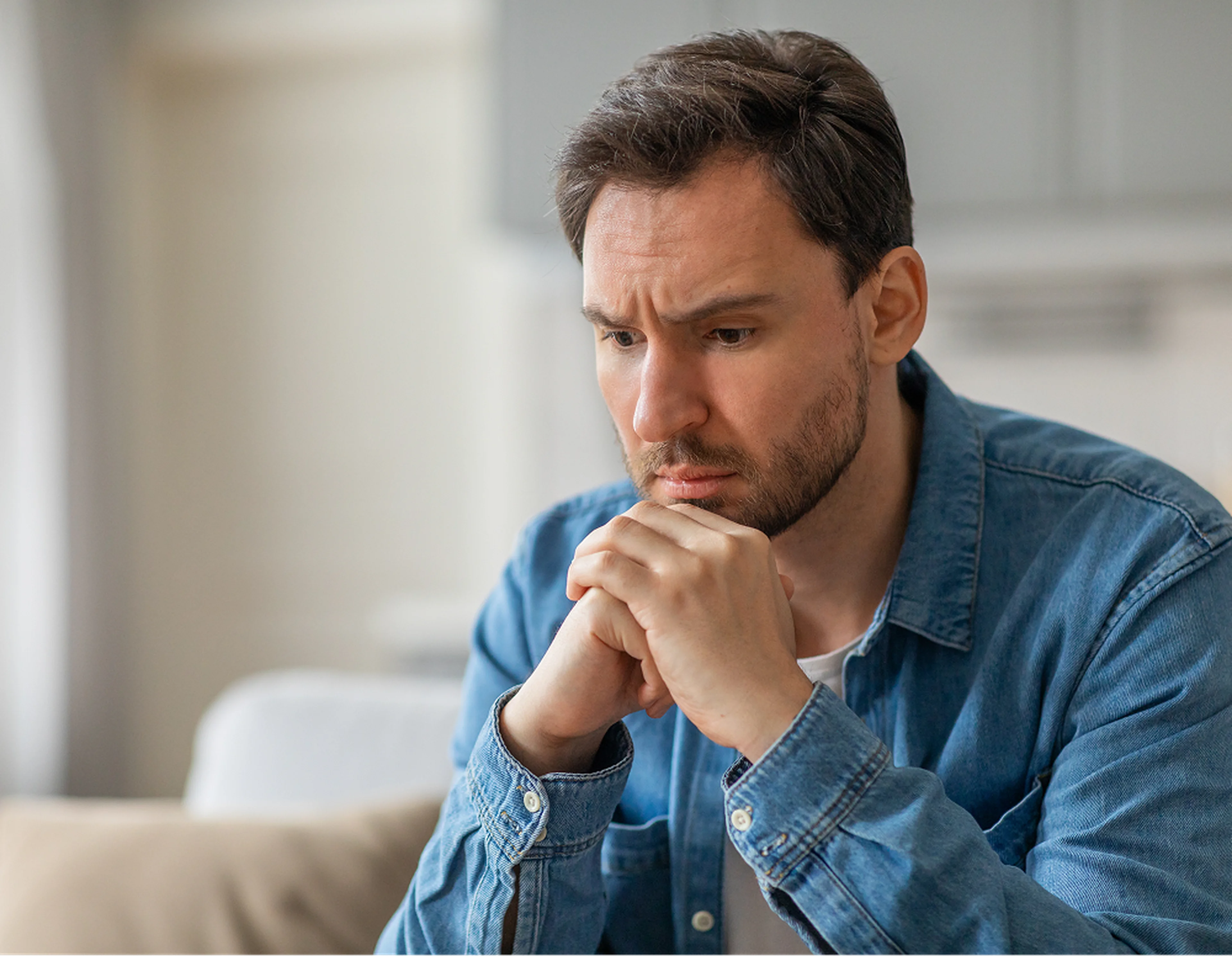 A middle-aged man in a blue denim shirt sits pensively with his hands clasped near his chin, appearing deep in thought in a softly lit indoor setting