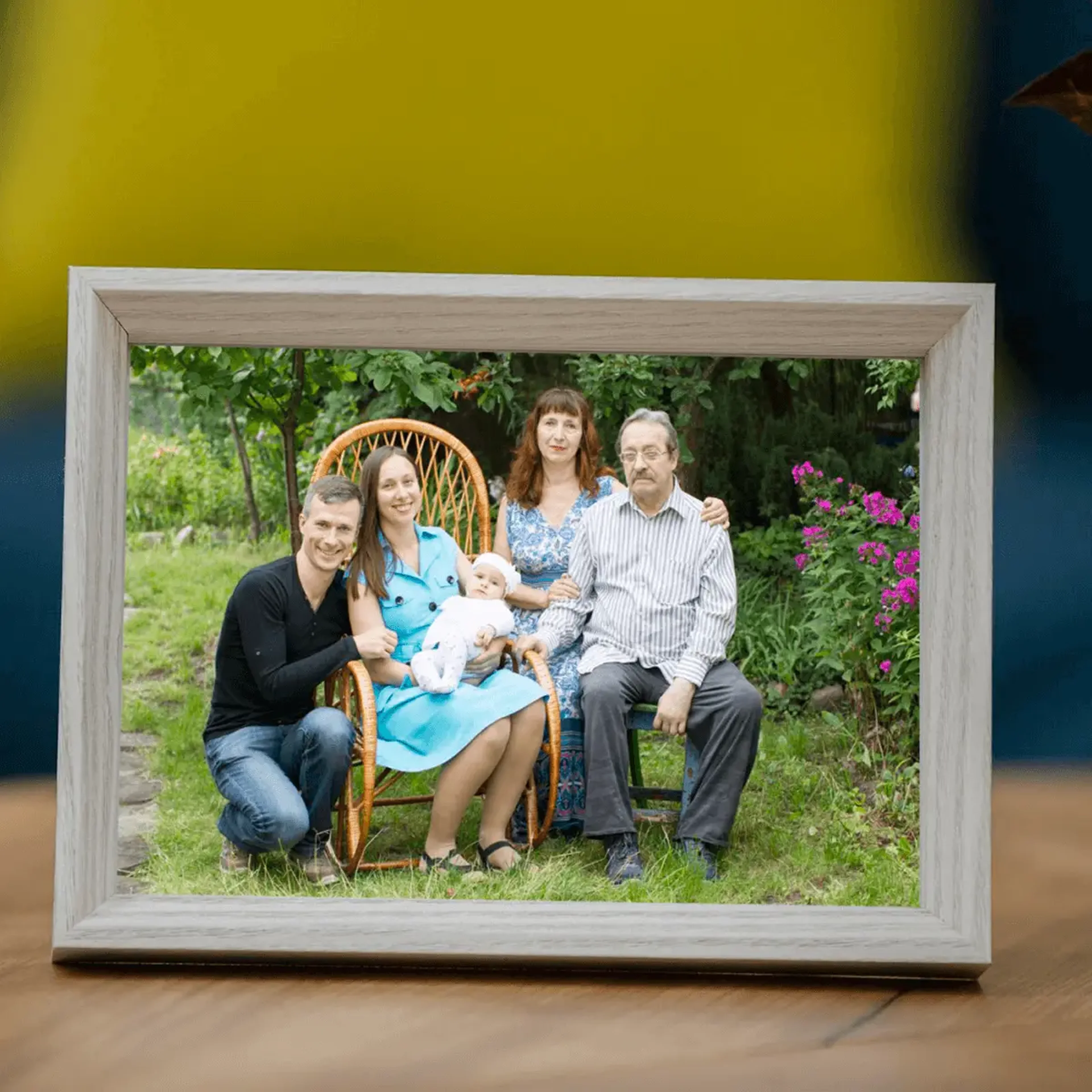 Framed family photo showing five people including parents with baby posed outdoors in wicker chair surrounded by greenery and flowers