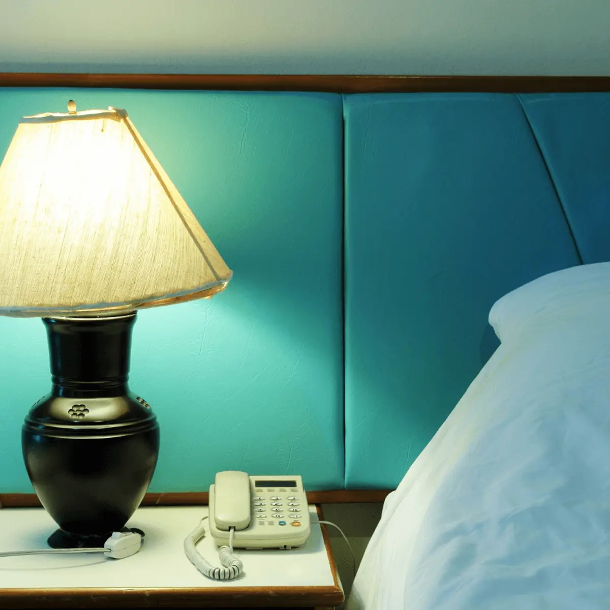 Bedside table in hotel room with ceramic table lamp, white telephone, and white bedding against teal upholstered headboard