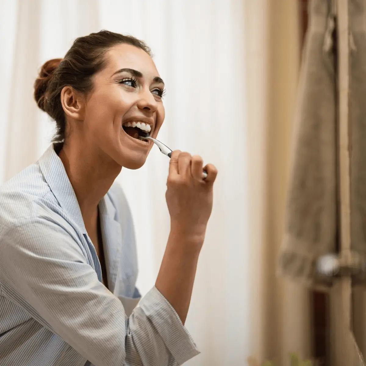 Smiling woman in light blue striped shirt brushing her teeth with toothbrush in bright bathroom