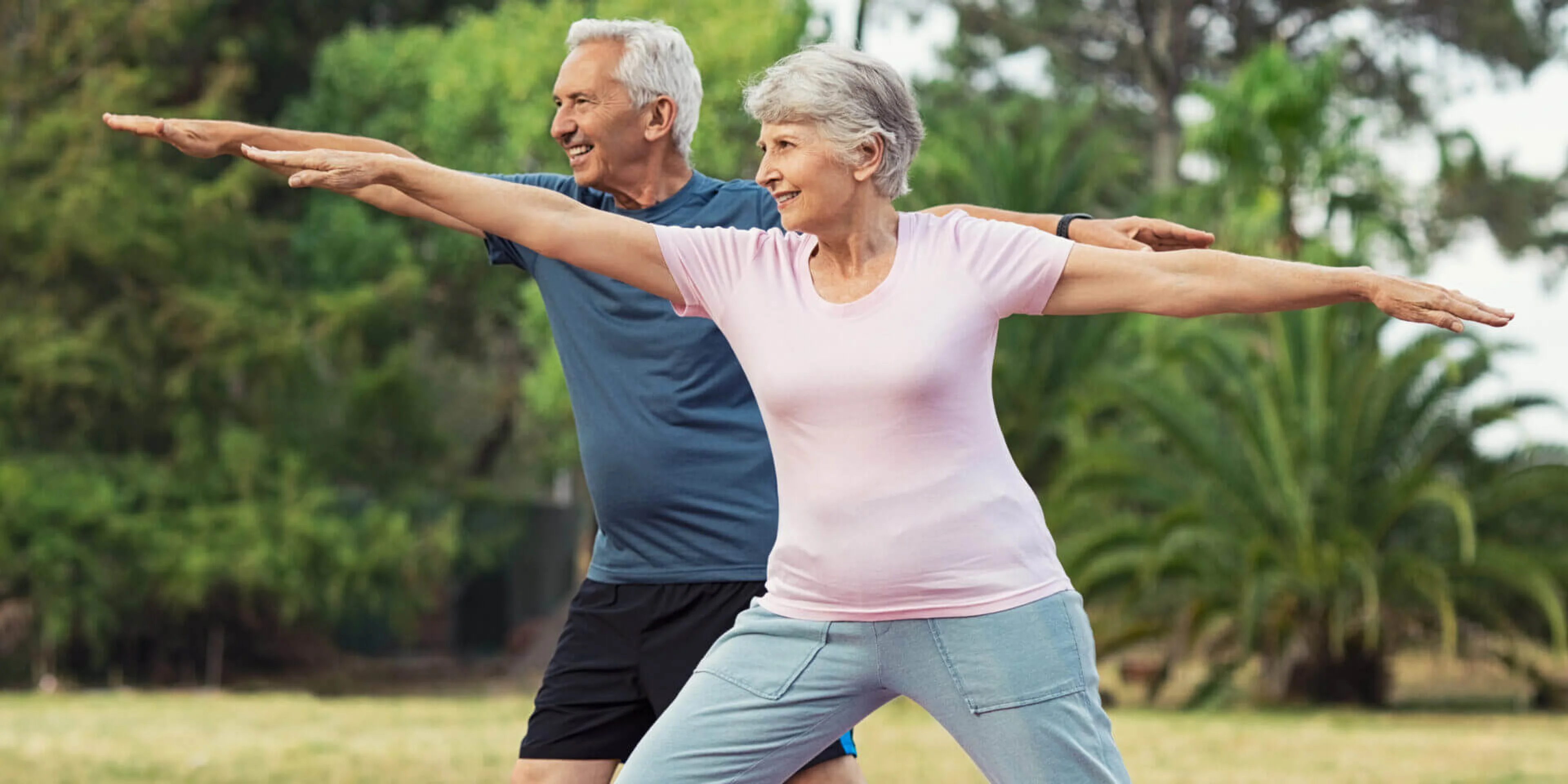 Two elderly people practice yoga outdoors, standing in a warrior pose with arms extended. They appear focused and relaxed, surrounded by greenery.