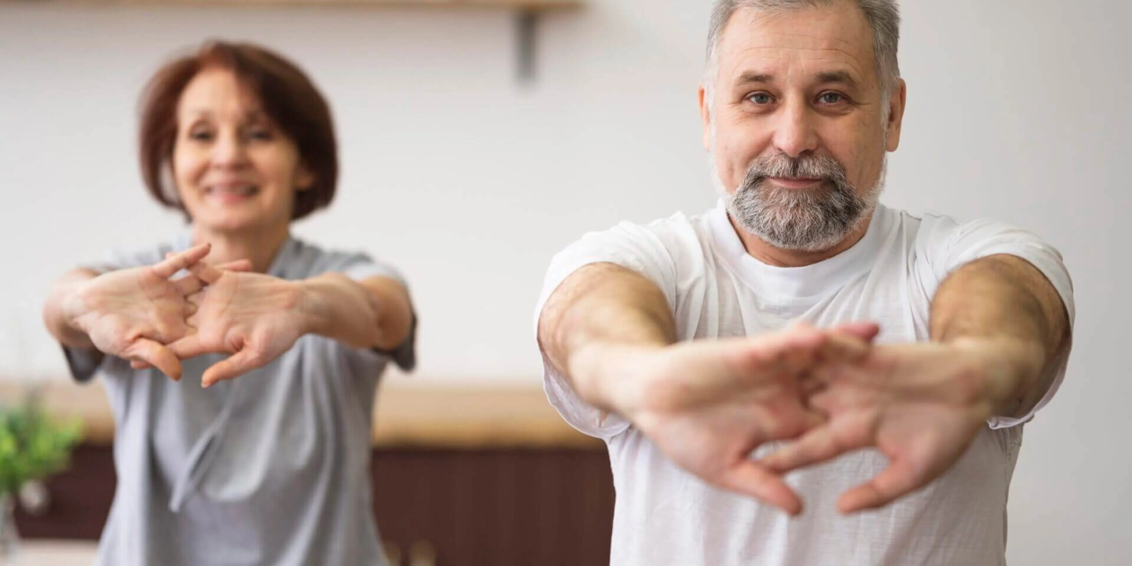 Older man and woman smiling, stretching their arms forward together in a bright indoor setting, wearing casual clothing.