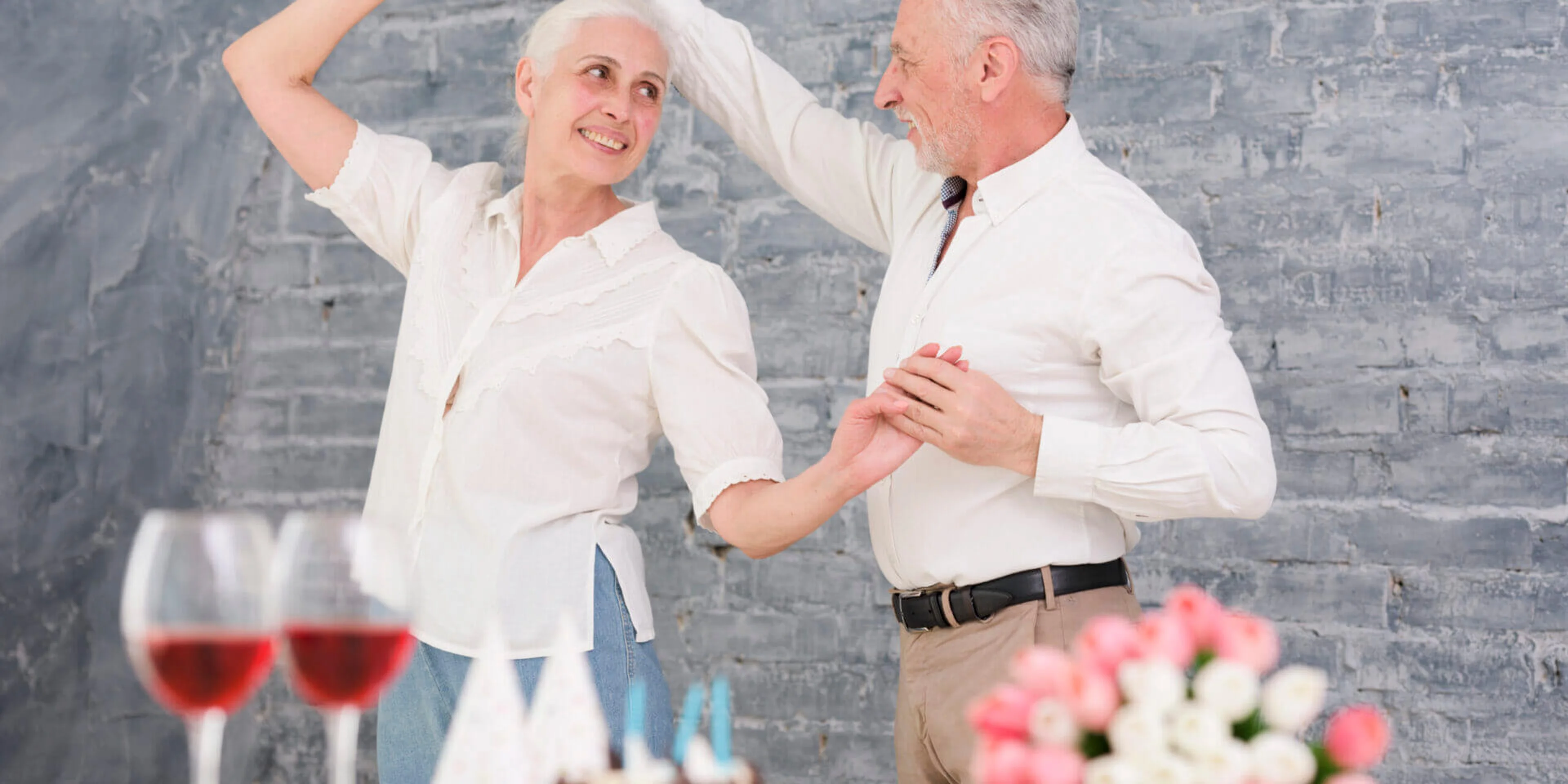Elderly couple dancing joyfully near a table with two wine glasses, birthday cake, and a bouquet of pink flowers against a gray brick wall.