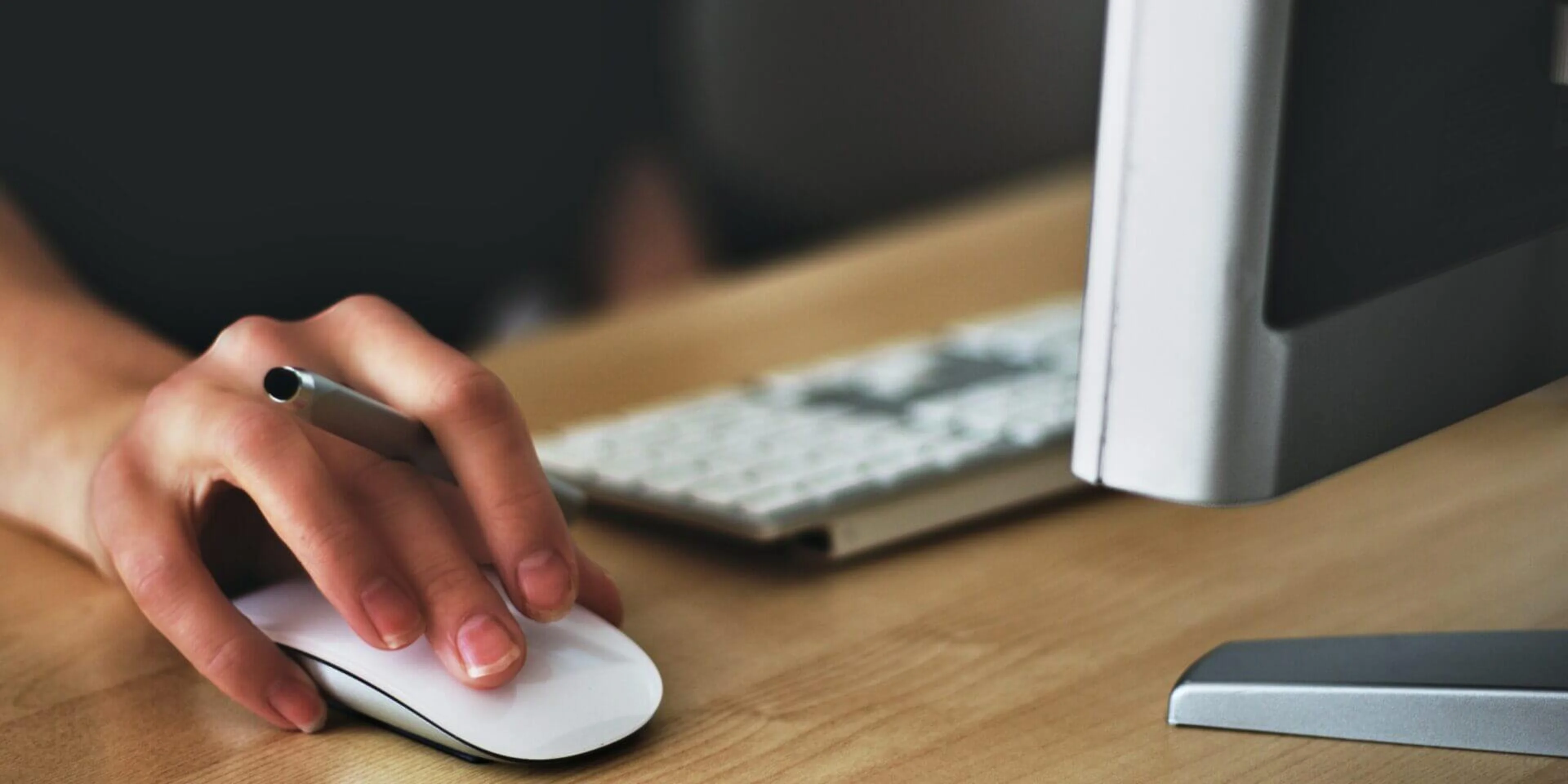 A person using a computer mouse with a pen resting on it beside a keyboard and monitor on a wooden desk.