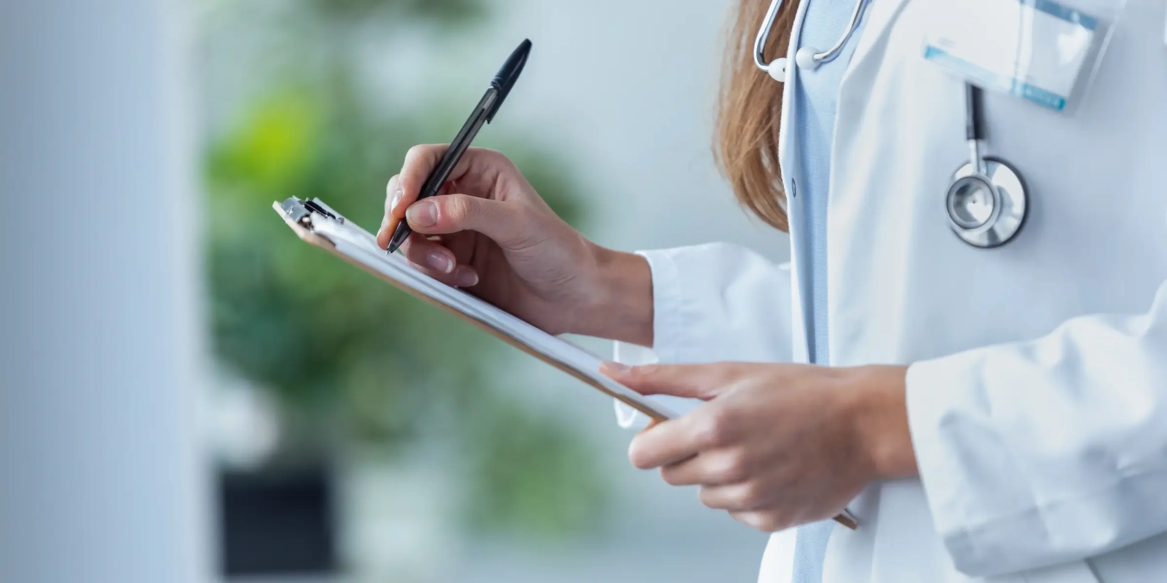 Female healthcare professional in white coat with stethoscope writing notes on clipboard in medical office