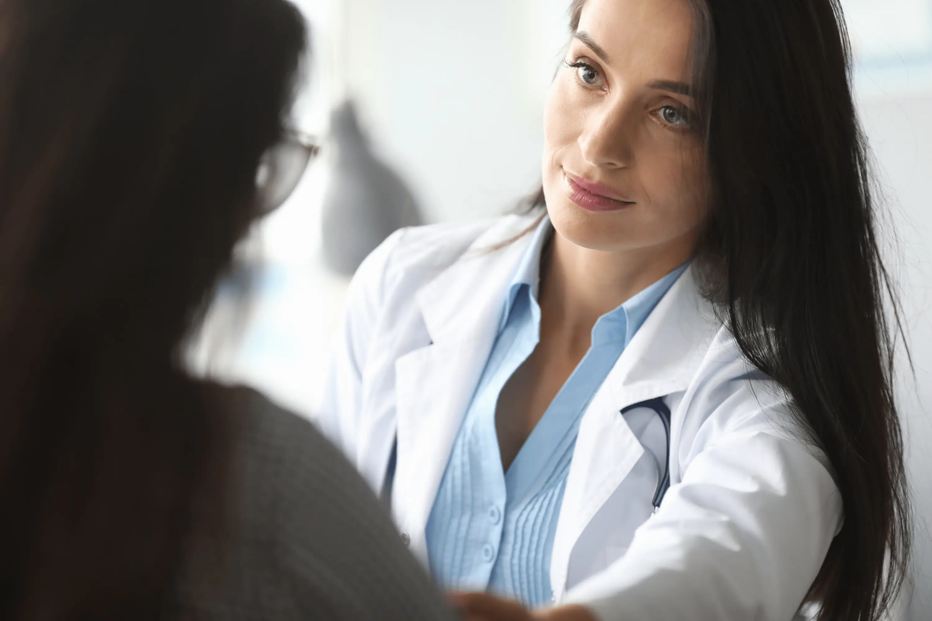 Female doctor in white coat and blue shirt listening attentively to patient during medical consultation in bright clinical setting