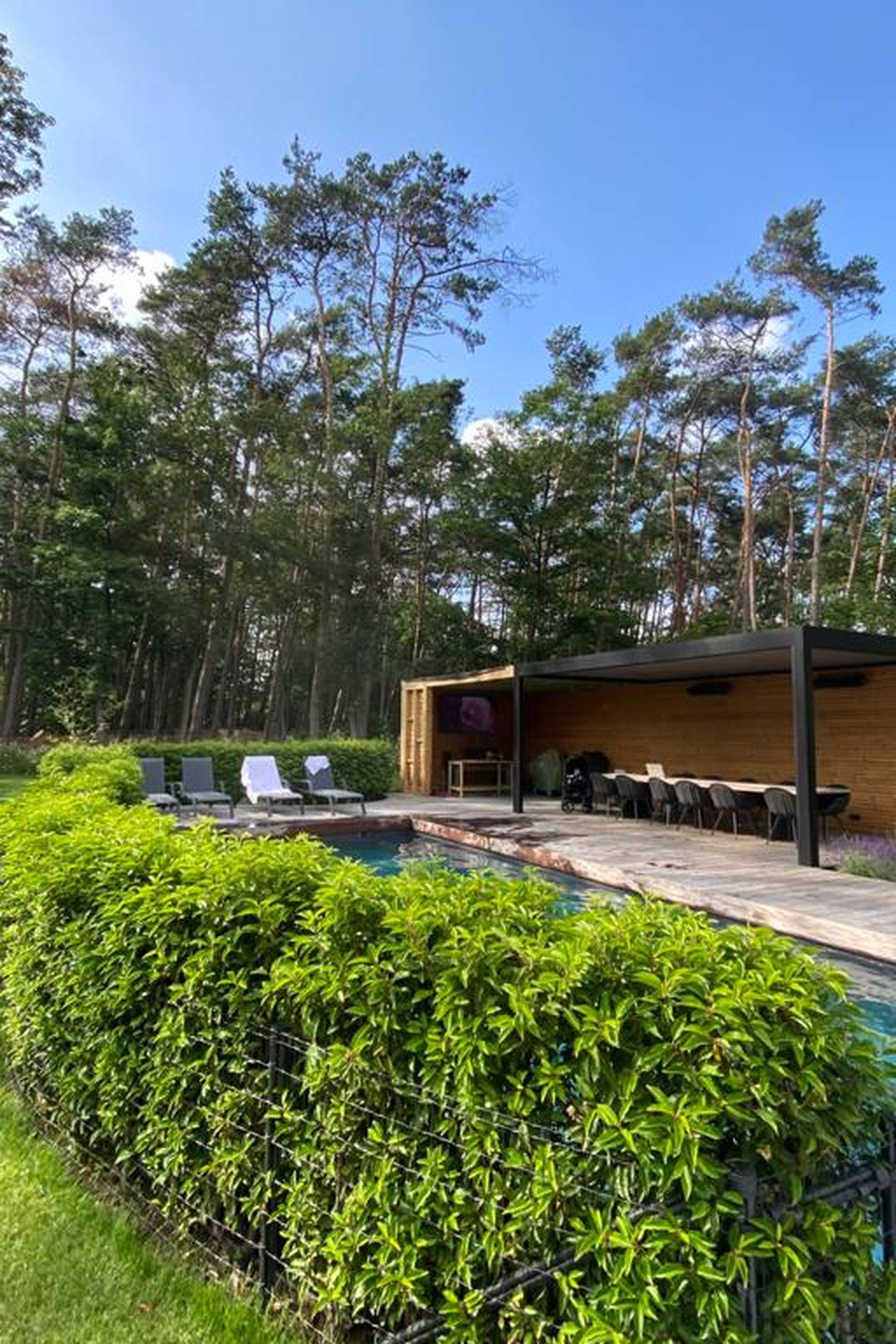 Ruime tuin met zwembad en een vrijstaande Brustor lamellenpergola, die een schaduwrijke eet‑ en loungezone vormt langs het water.