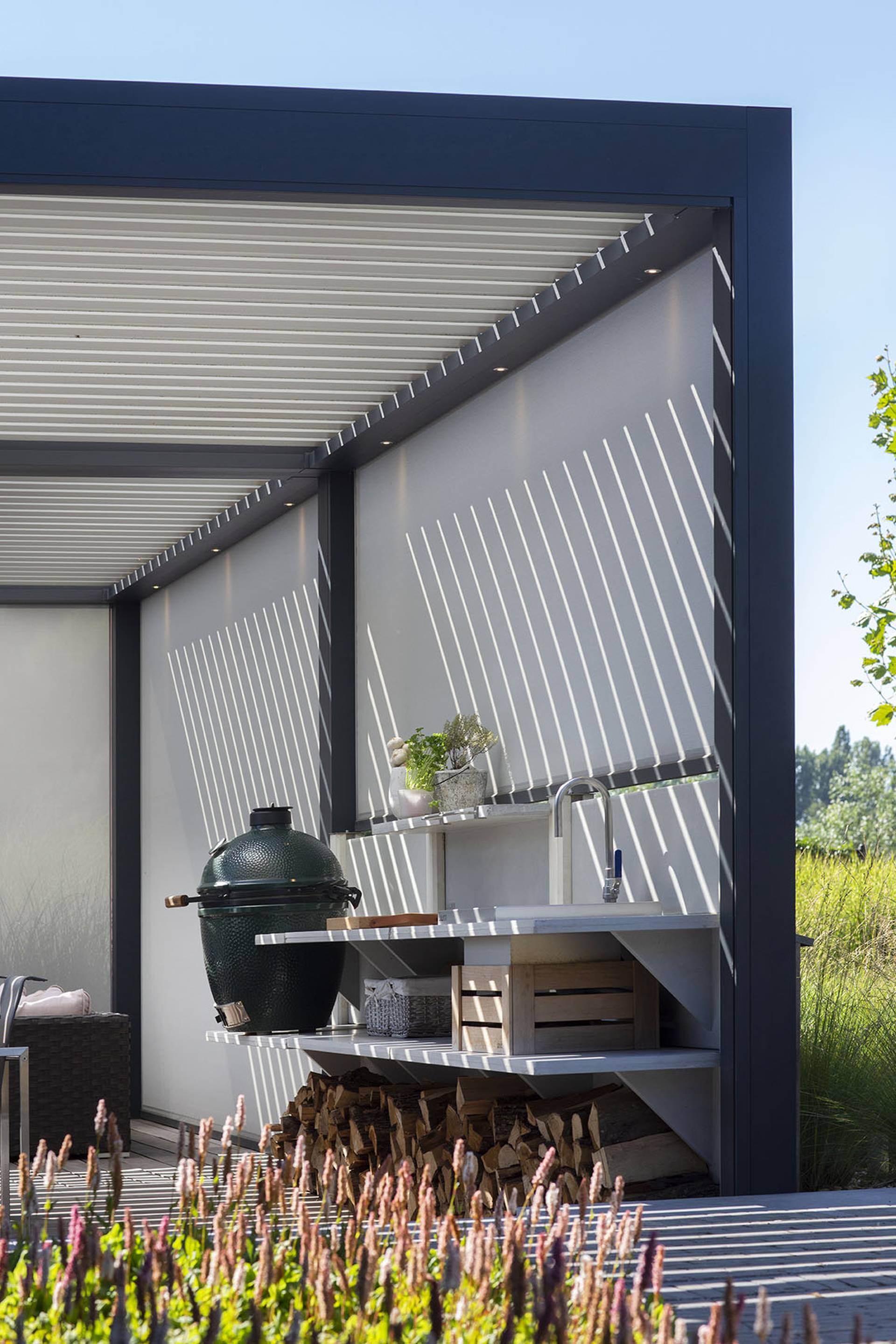 Outdoor kitchen area under an aluminium pergola with tilting louvred roof and lowered ZIP‑screens creating a striped light pattern on the wall.