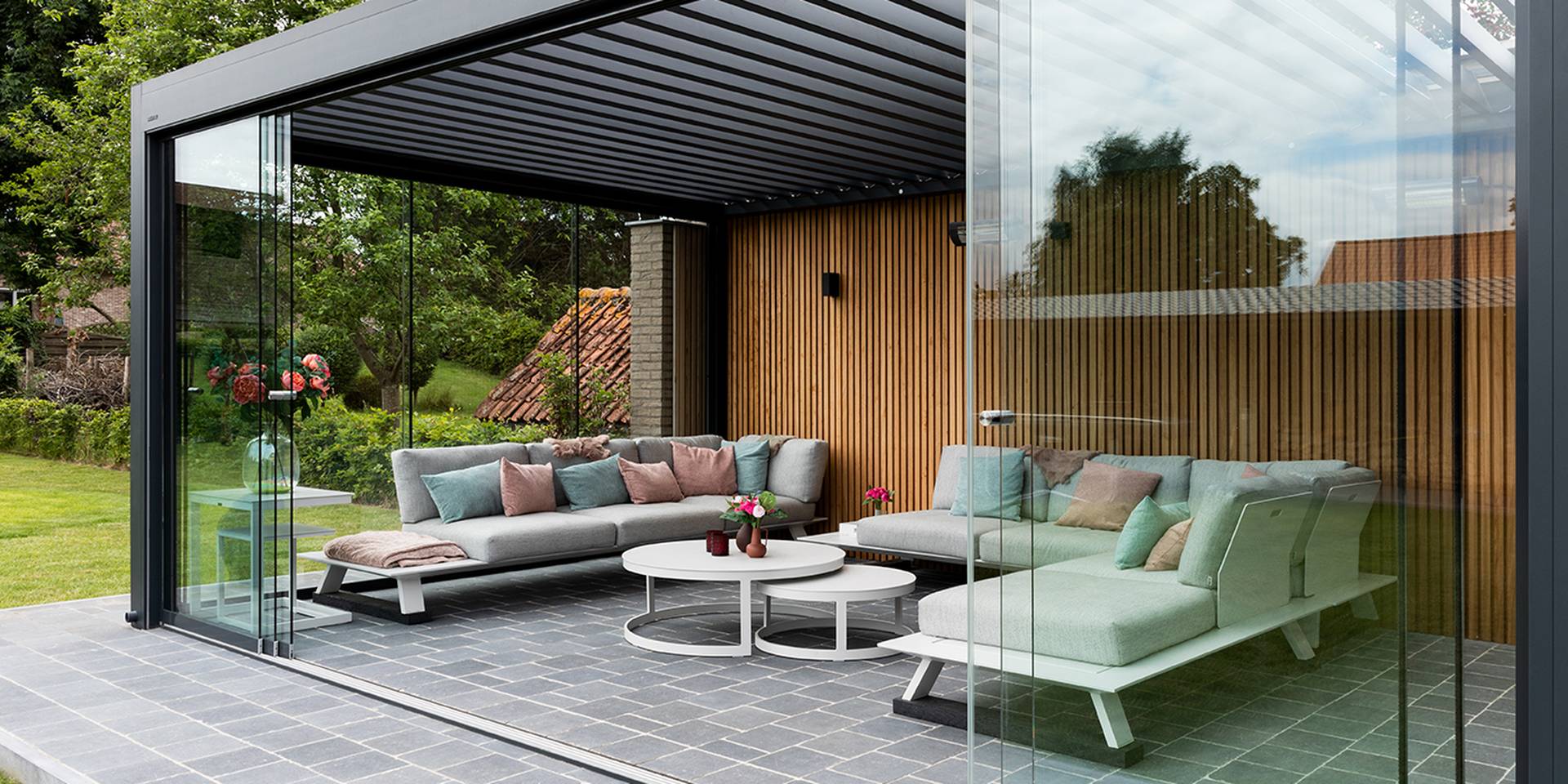Aluminium pergola with tilting louvred roof and glass sliding walls enclosing an outdoor lounge area with wooden wall cladding.