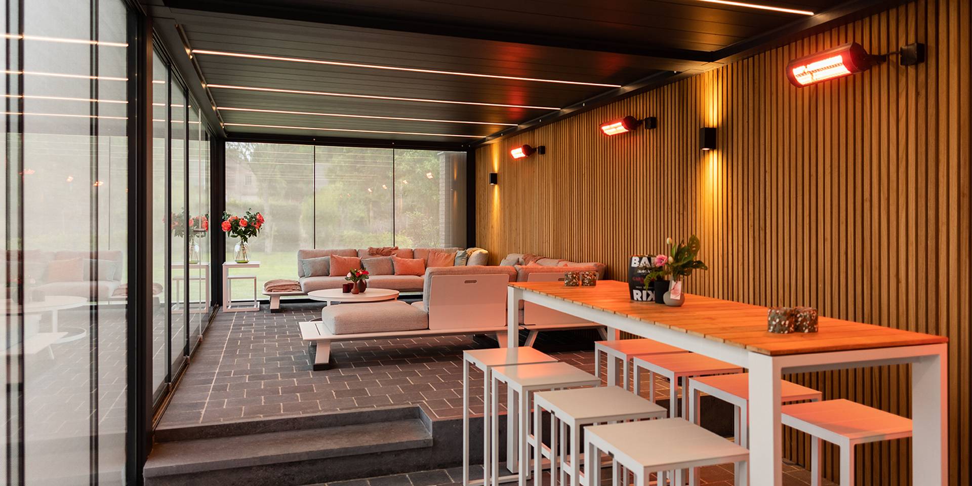 Aluminium pergola with tilting louvred roof and glass sliding walls, featuring integrated LED lighting, outdoor bar table and lounge seating.