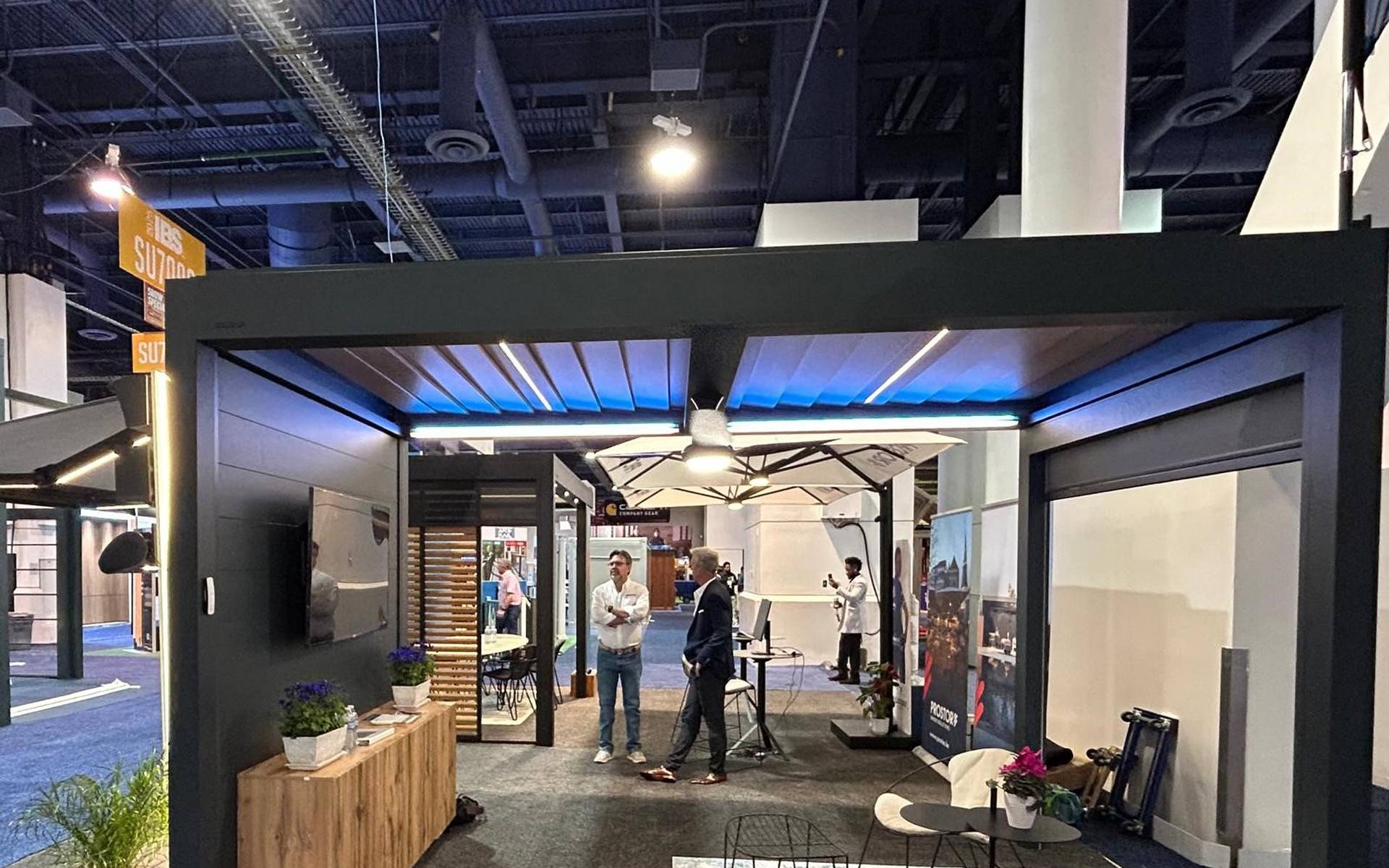 Brustor exhibition booth showcasing an aluminium pergola with integrated lighting, outdoor furniture and product displays at an indoor trade fair.