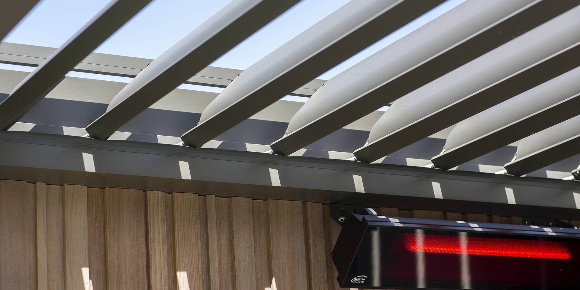 Gros plan d’une pergola en aluminium à lames orientables, laissant filtrer la lumière, avec chauffage de terrasse intégré en dessous.