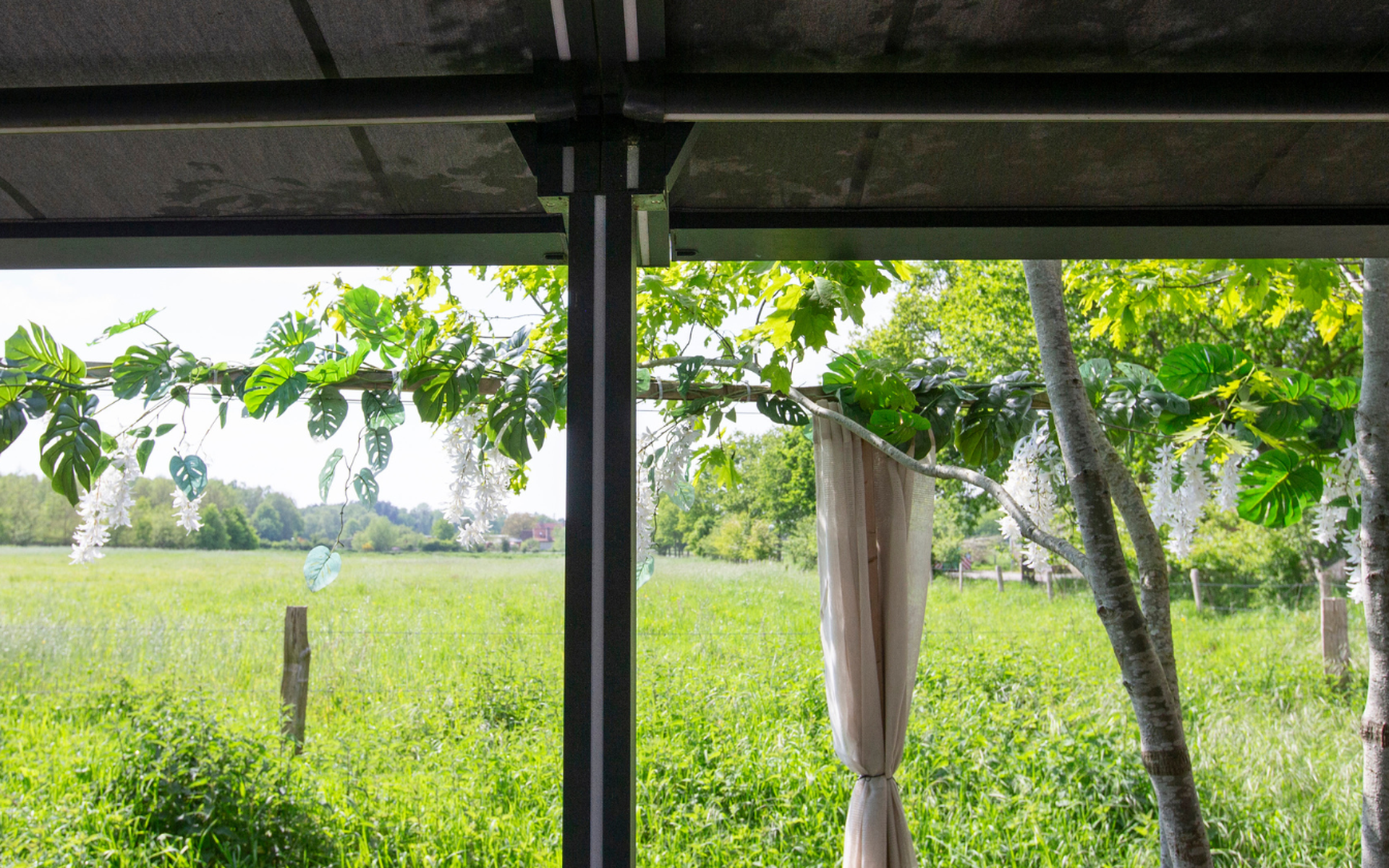 Blick unter einer Aluminium-Pergola auf eine grüne Landschaft mit dekorativem Laub und Bäumen.
