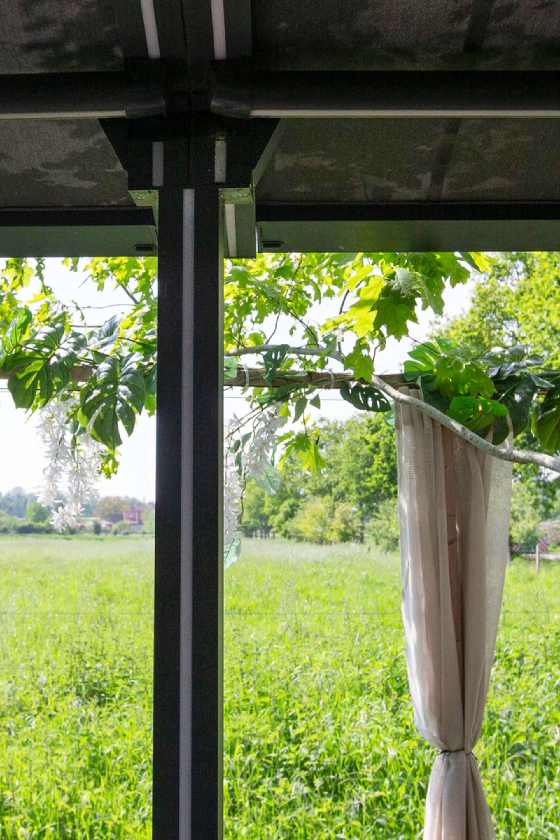 Zicht van onder een aluminium pergola op een groen landschap met decoratieve beplanting en bomen.