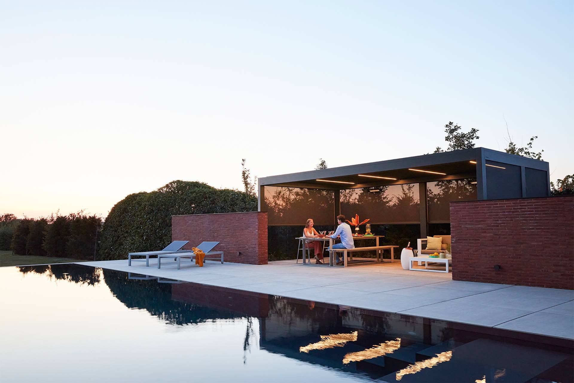 Abendliche Ess‑ und Loungezone am Pool unter einer Brustor B200(XL) Lamellenpergola mit integrierten ZIP‑Screens und warmem LED‑Licht für eine gemütliche Atmosphäre.