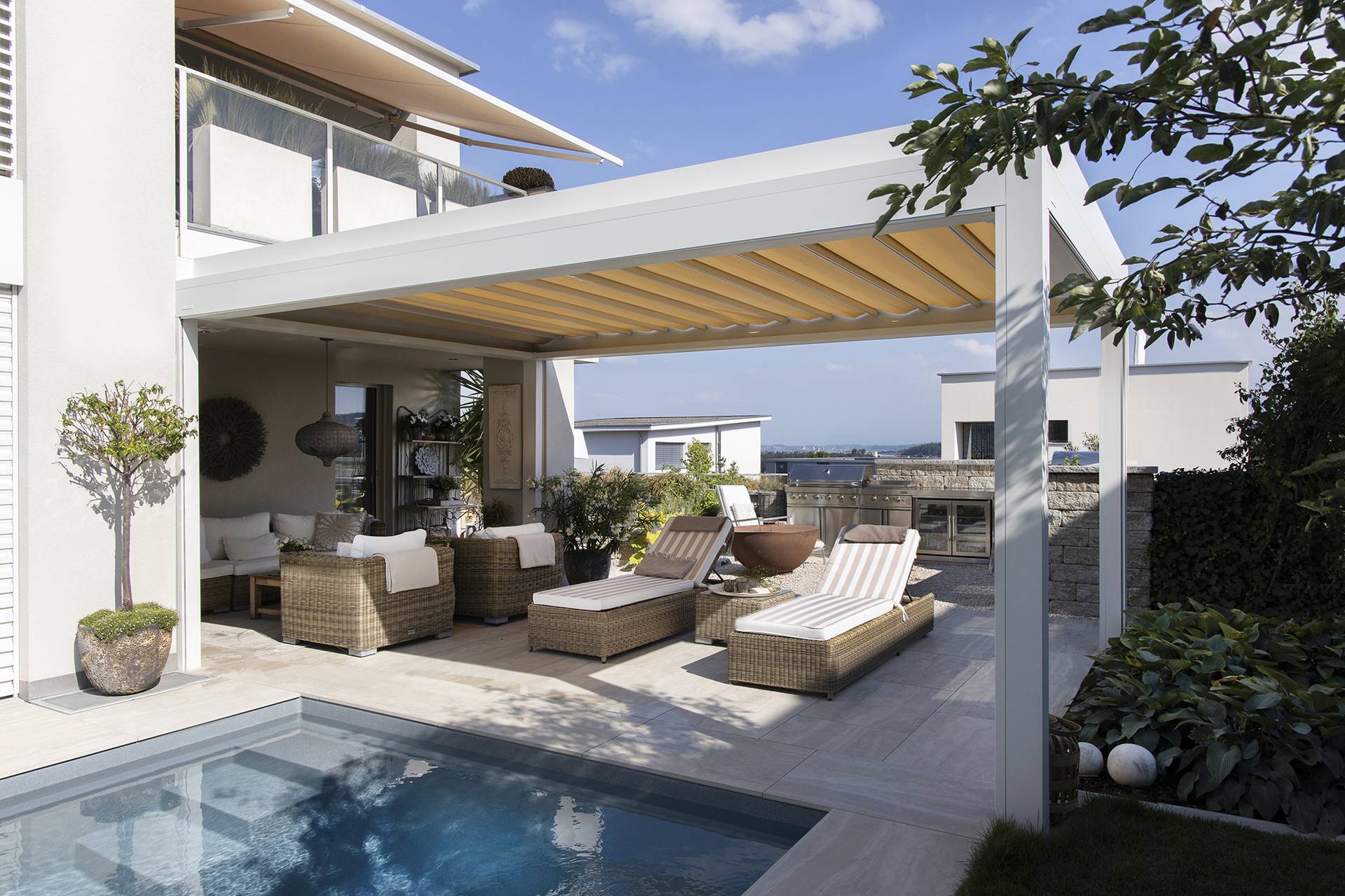 Aluminium pergola with a retractable fabric roof shading an outdoor lounge area with sunbeds beside a private pool.