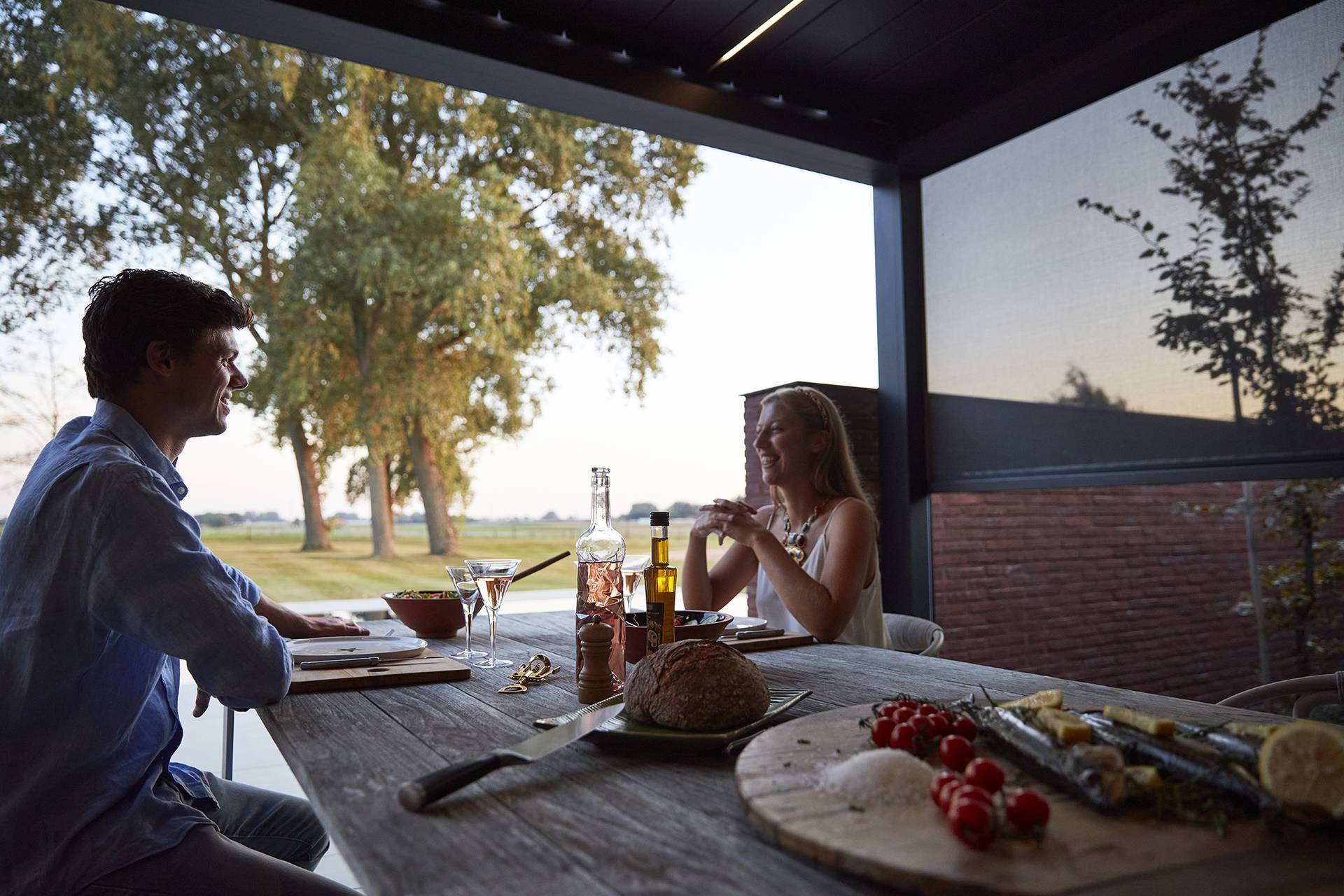 Abendliche Essensszene unter einer Brustor B200(XL) Lamellenpergola mit warmem LED‑Licht, mit gedecktem Tisch und Blick auf den Garten.