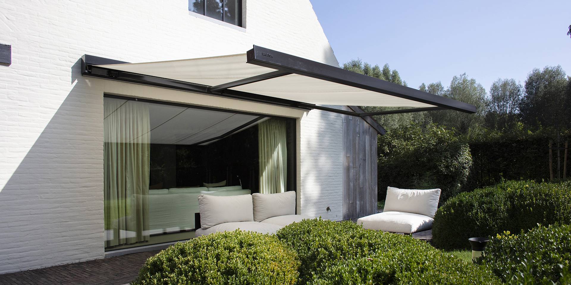 Store banne moderne en aluminium offrant de l’ombre à une terrasse avec mobilier lounge, installé au‑dessus de grandes baies vitrées donnant sur un jardin paysager.