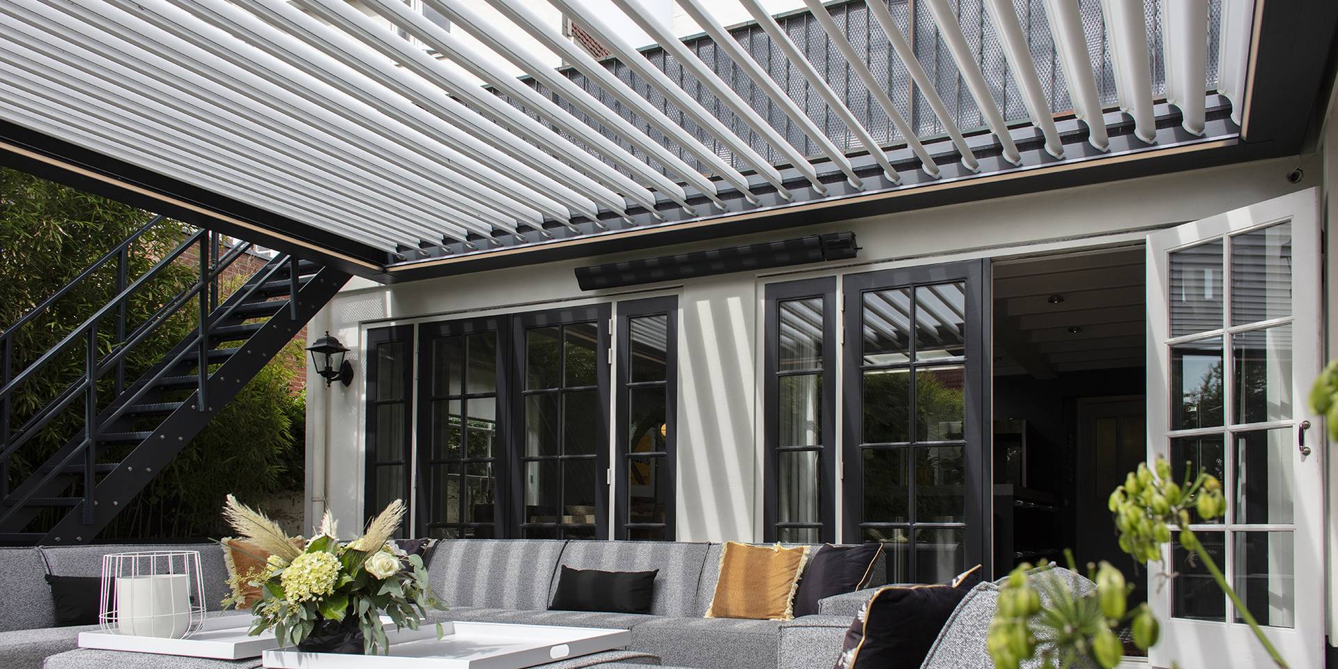 Aluminium pergola with tilting louvred roof attached to a house, creating a shaded outdoor lounge area beside large glass doors.