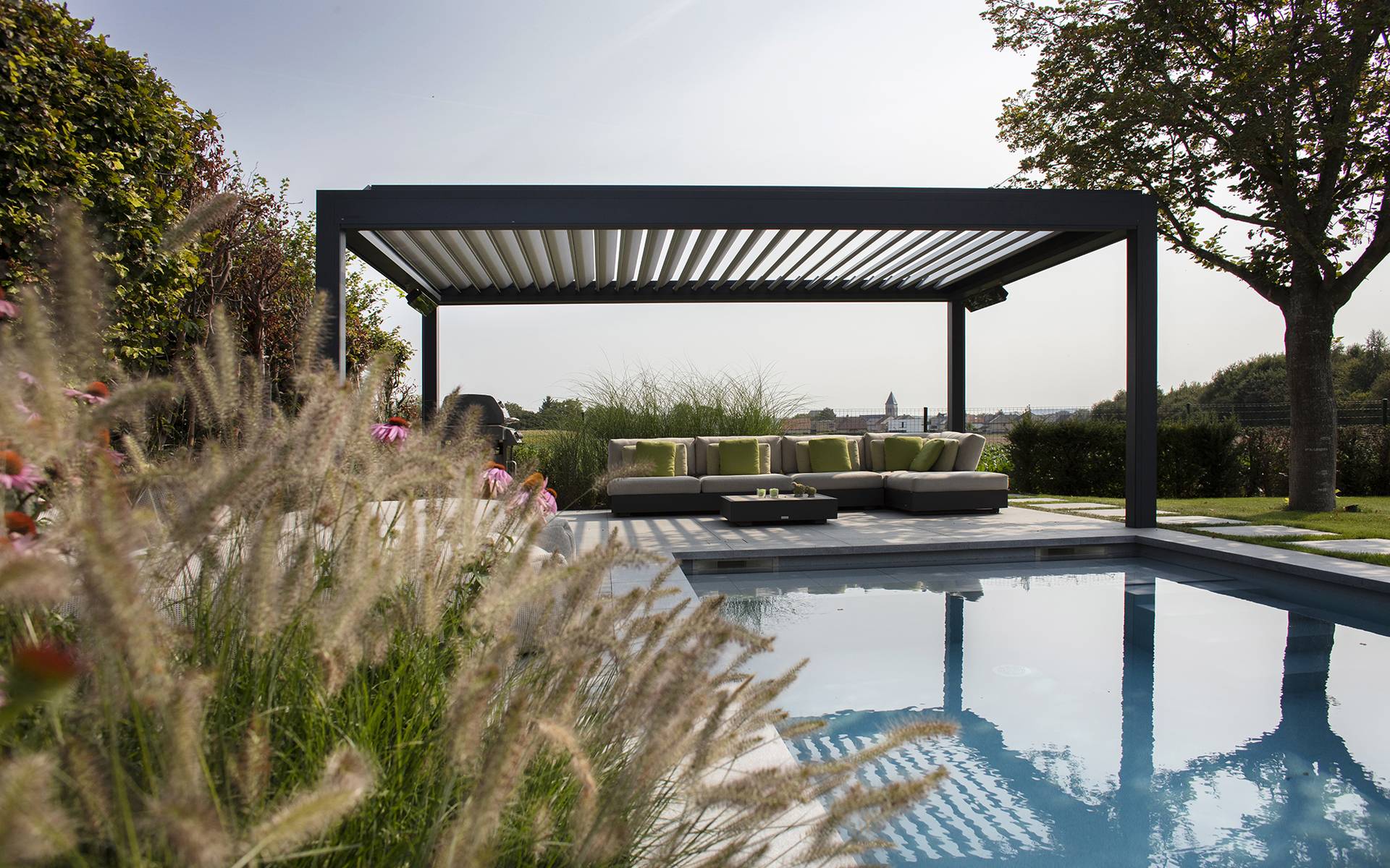 Aluminium pergola with tilting louvred roof installed beside a swimming pool, shading an outdoor lounge area in a landscaped garden.