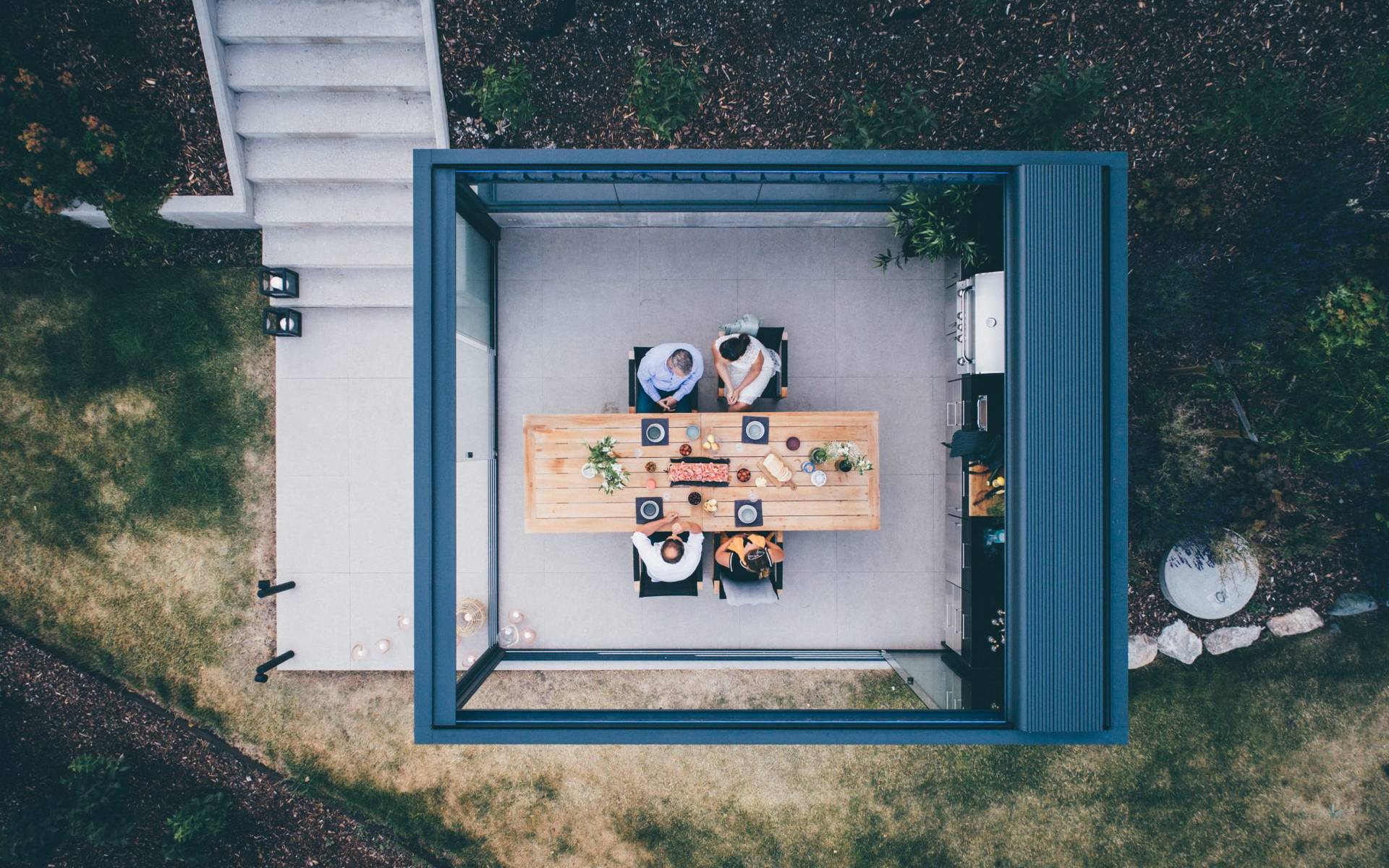 Aerial view of an aluminium pergola with retractable roof shading an outdoor dining area on a tiled terrace in a landscaped garden.