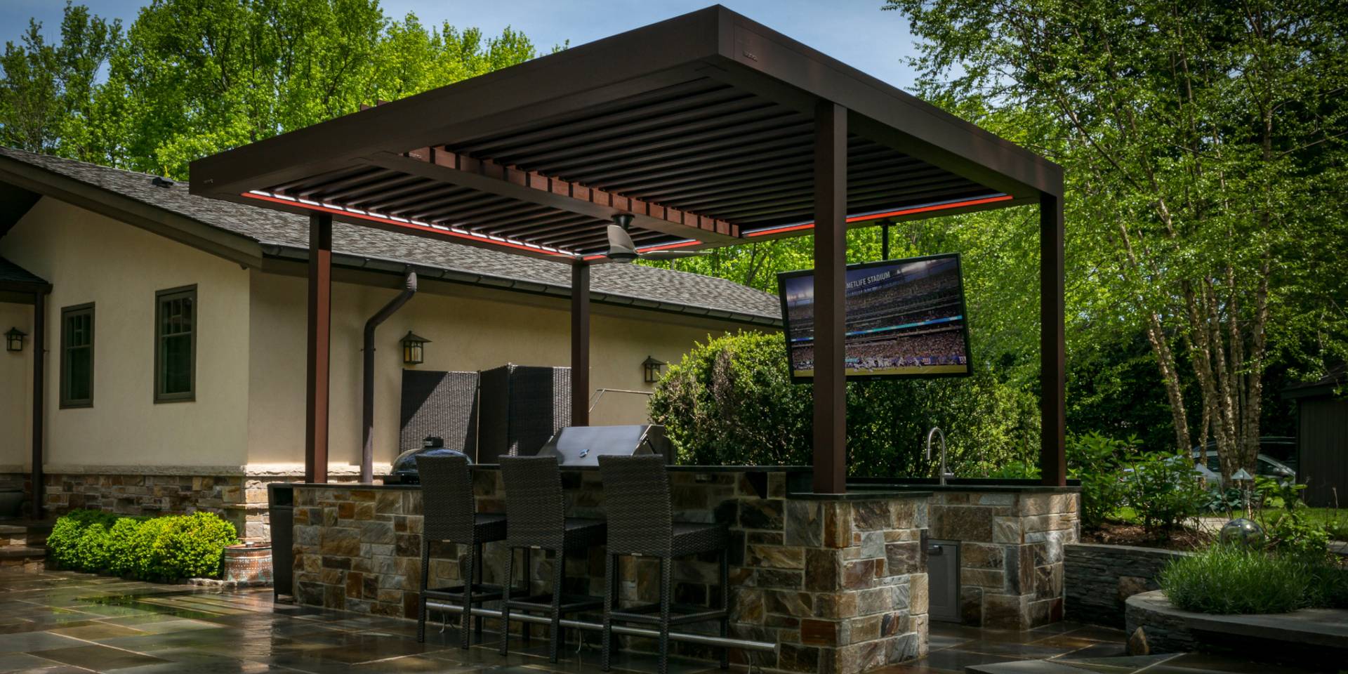 Outdoor bar area with a modern aluminium pergola featuring tilting louvres, integrated lighting and a mounted TV, set in a landscaped garden.