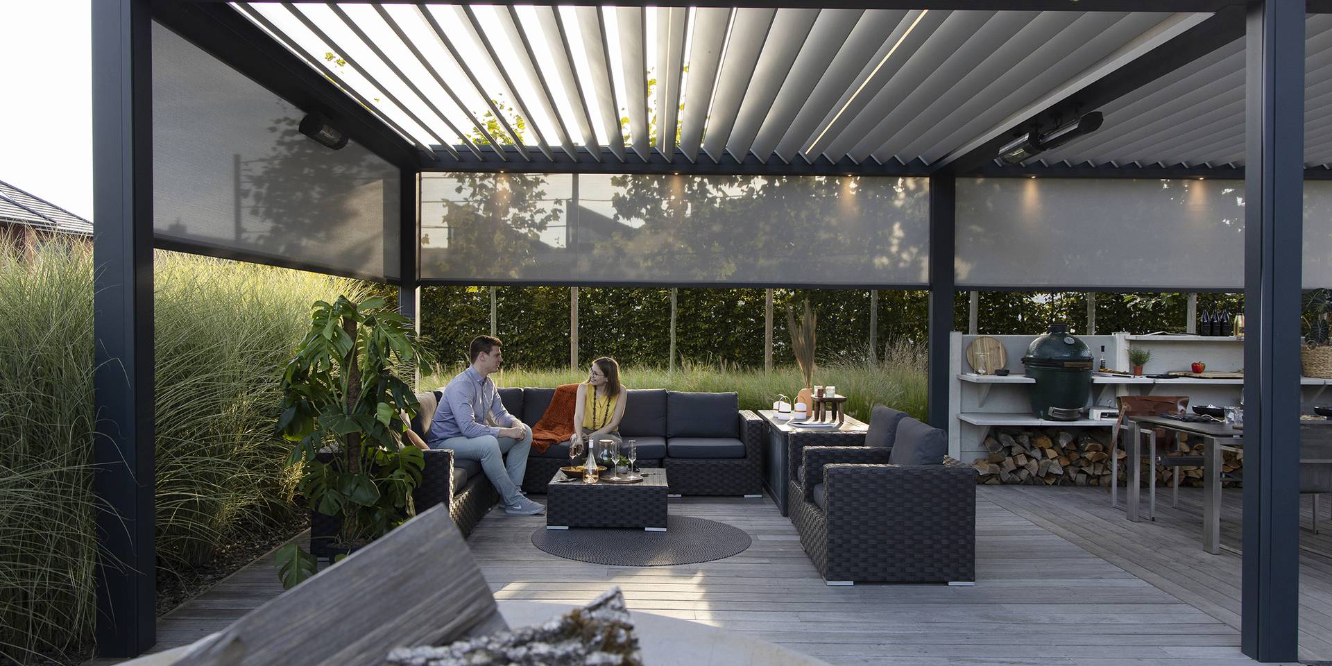 Aluminium pergola with tilting louvred roof and partially closed ZIP‑screens, shading an outdoor lounge and kitchen area on a wooden terrace.