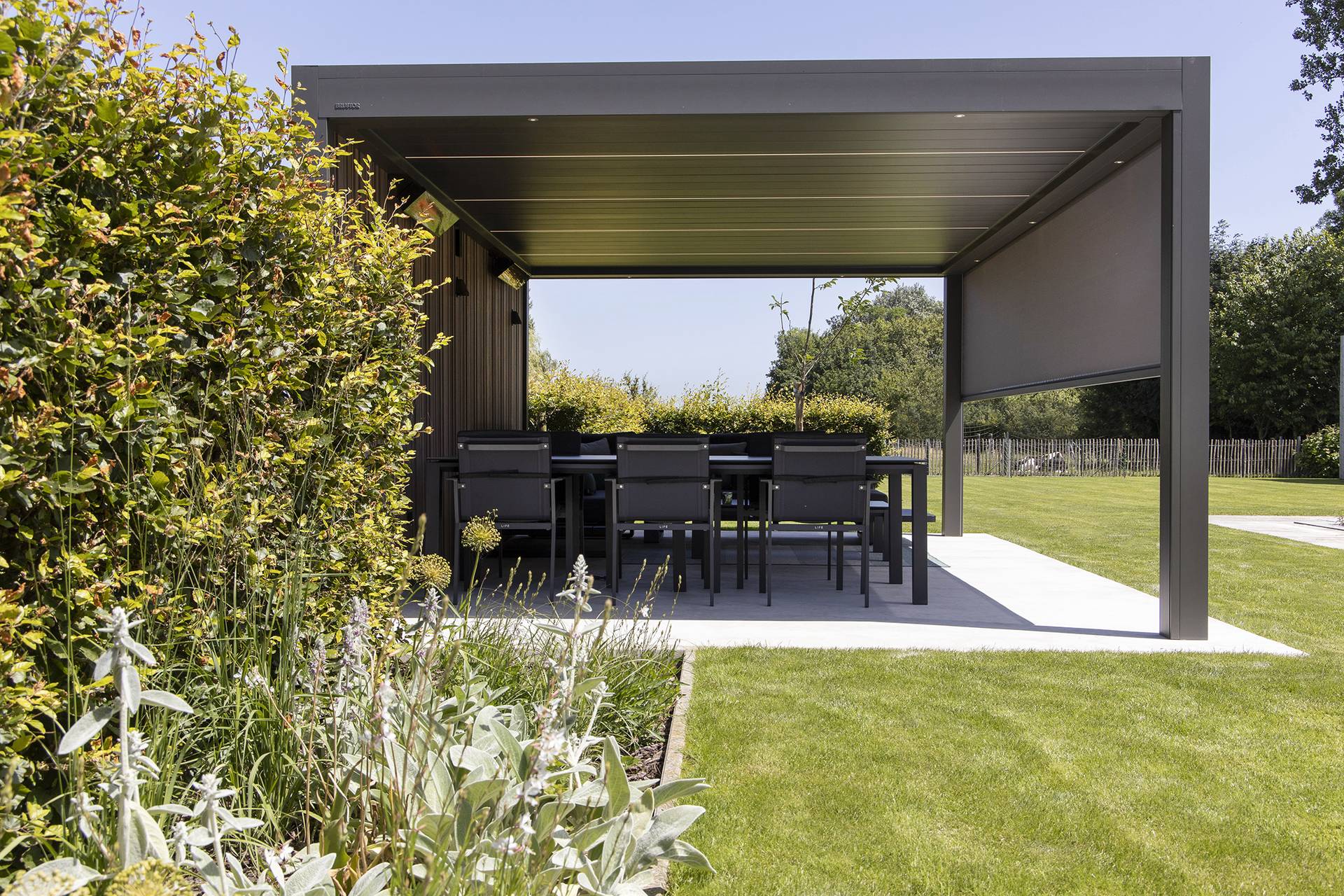 Outdoor dining area beneath an aluminium pergola with a solid roof, overlooking a landscaped garden.